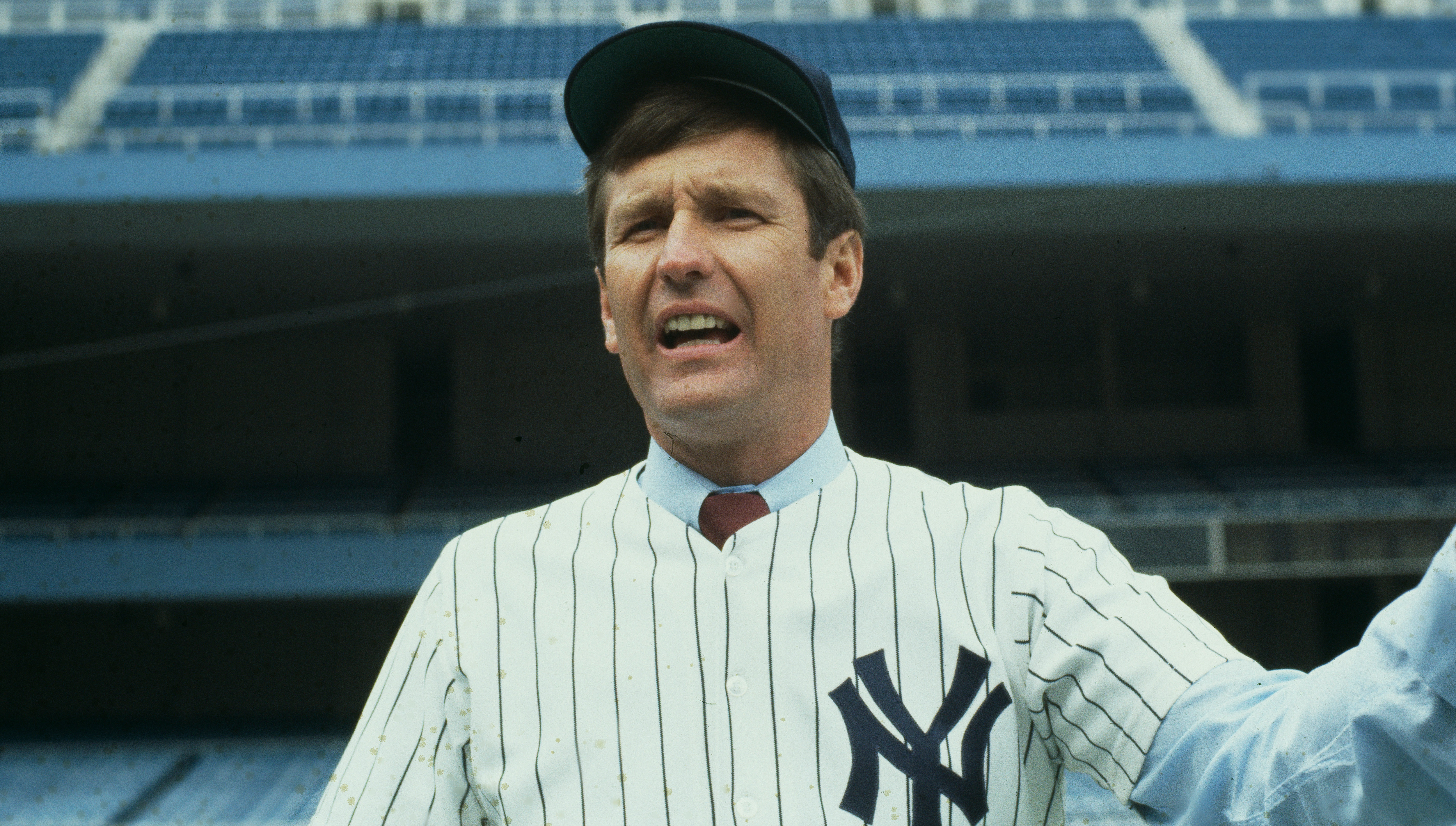 Tommy John in a New York Yankees uniform and cap is holding a baseball in a stadium.
