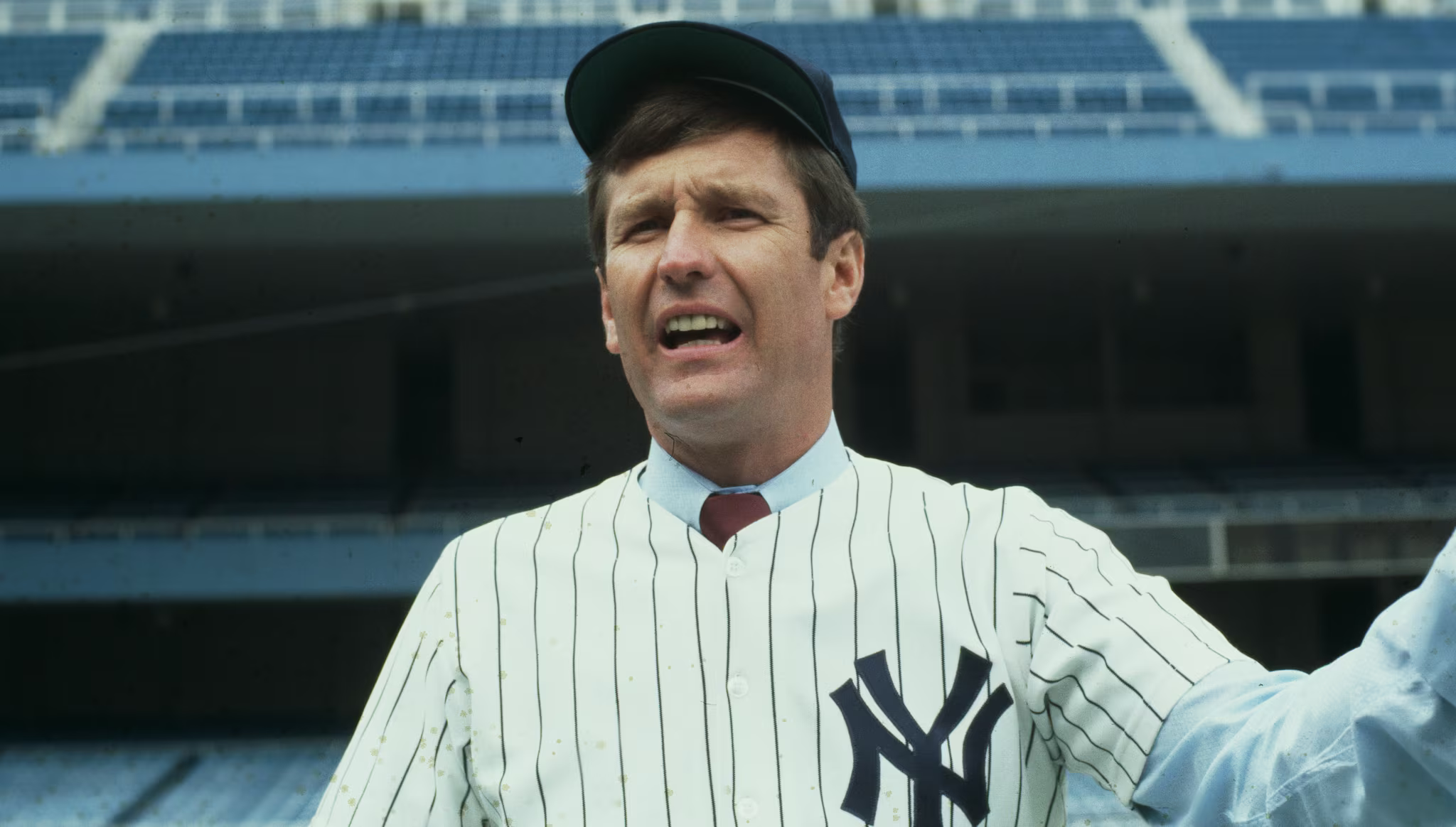 Tommy John in a New York Yankees uniform and cap is holding a baseball in a stadium.