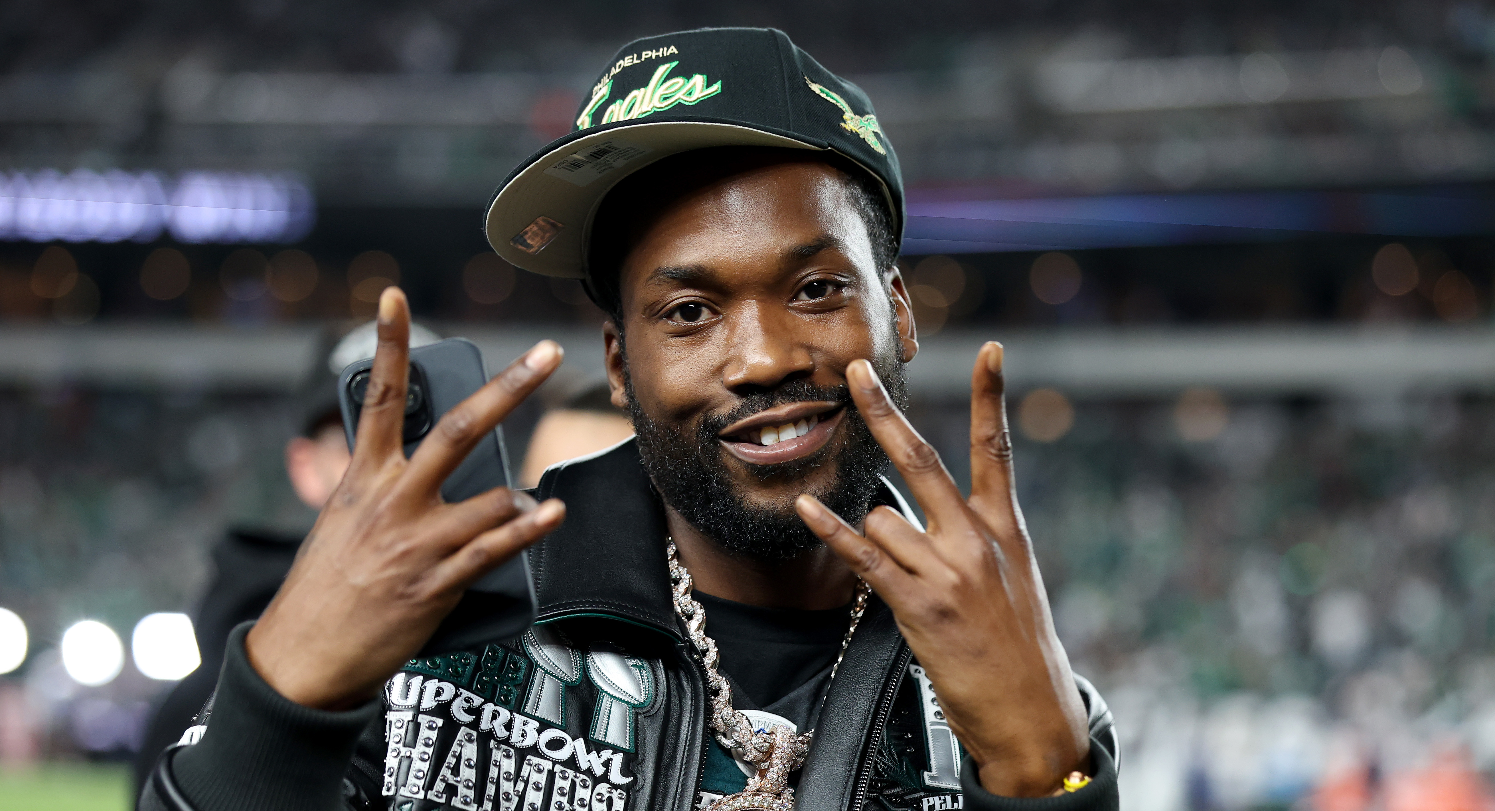Meeks Mill in Philadelphia Eagles cap and jacket, smiling and making hand gestures. The background is a stadium.