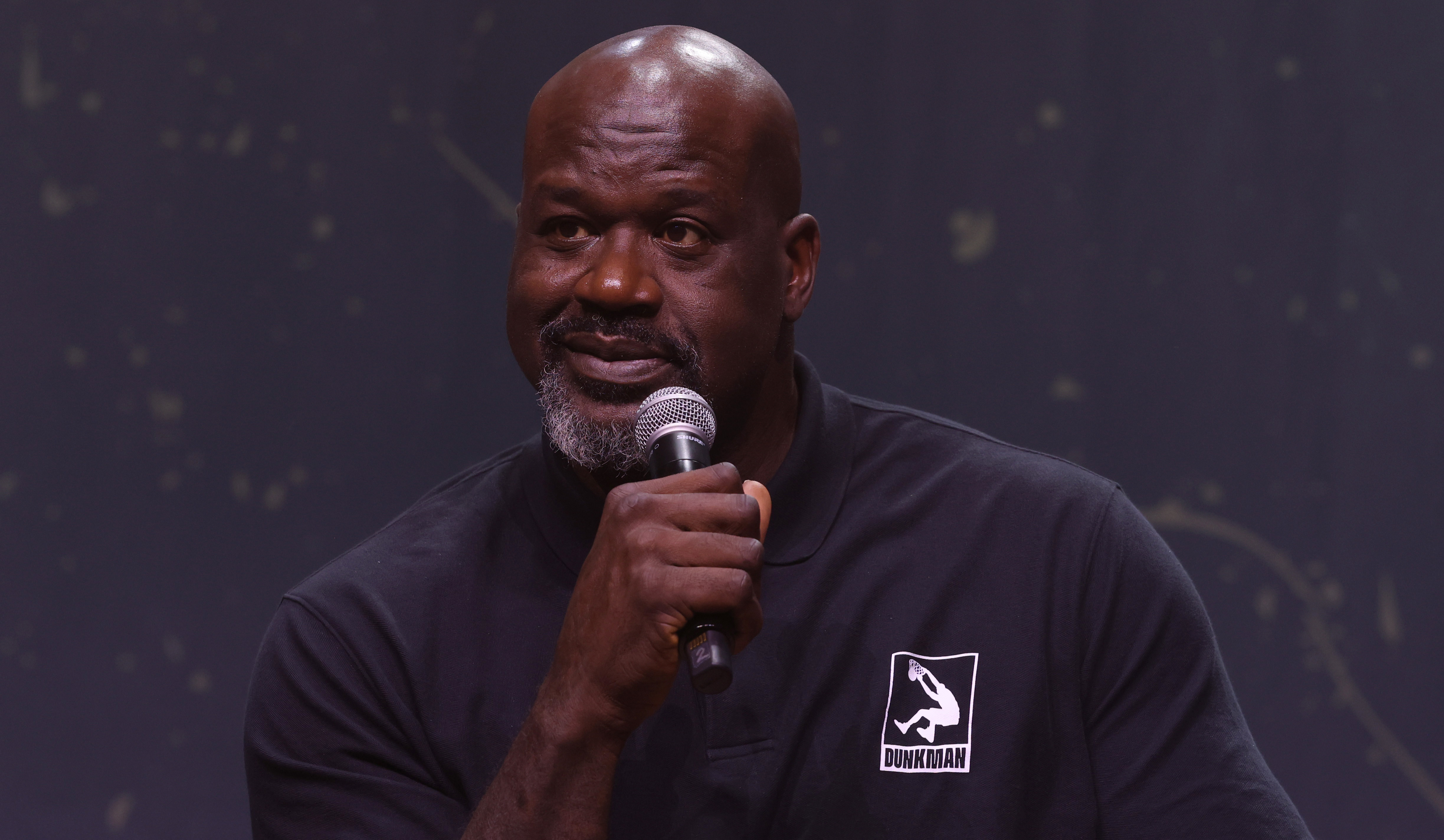 Shaquille O'Neal, wearing a black "Dunkman" polo, holds a microphone while speaking on stage.