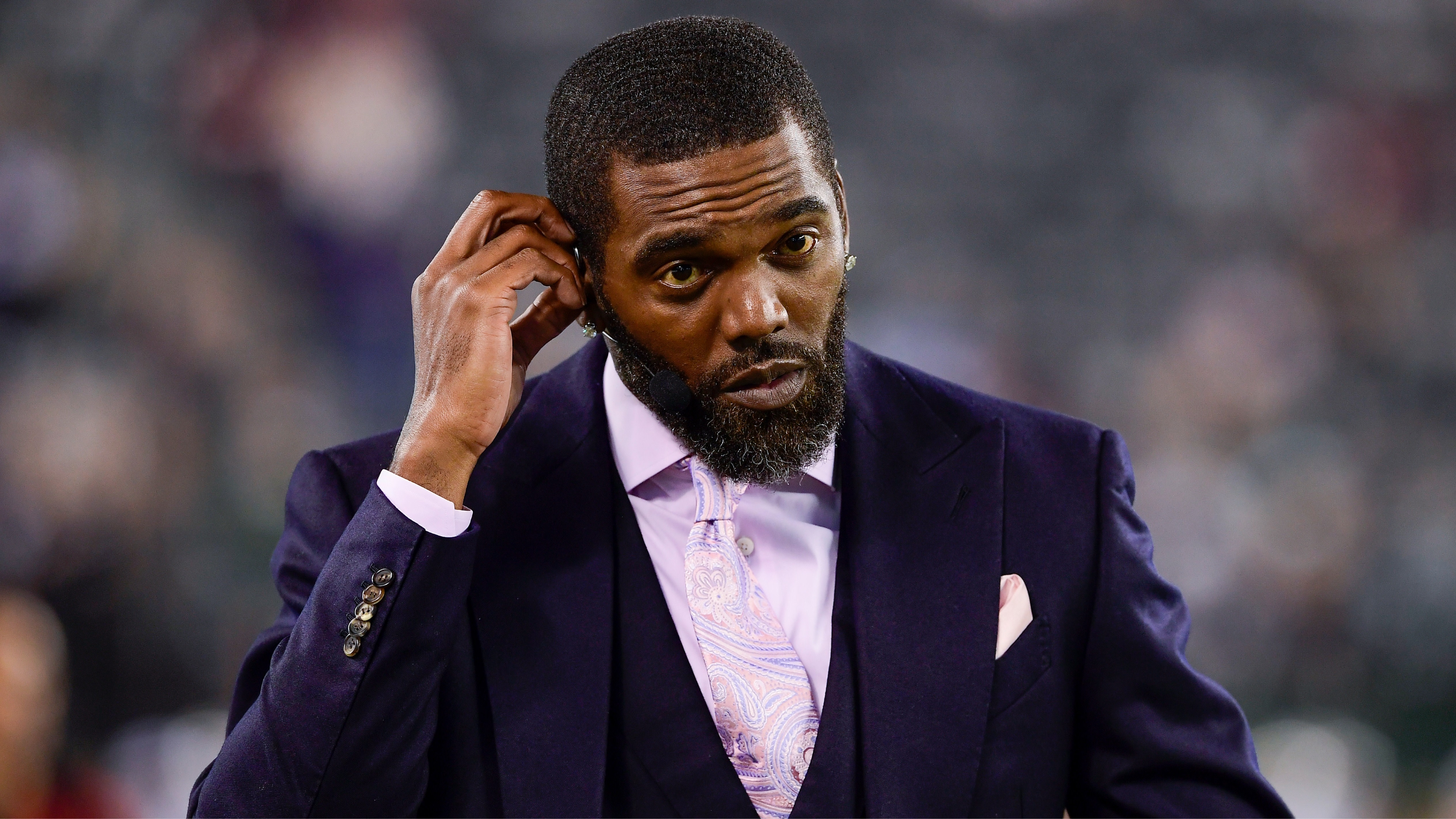 Randy Moss in a dark suit and light pink tie stands outdoors, touching his ear. 