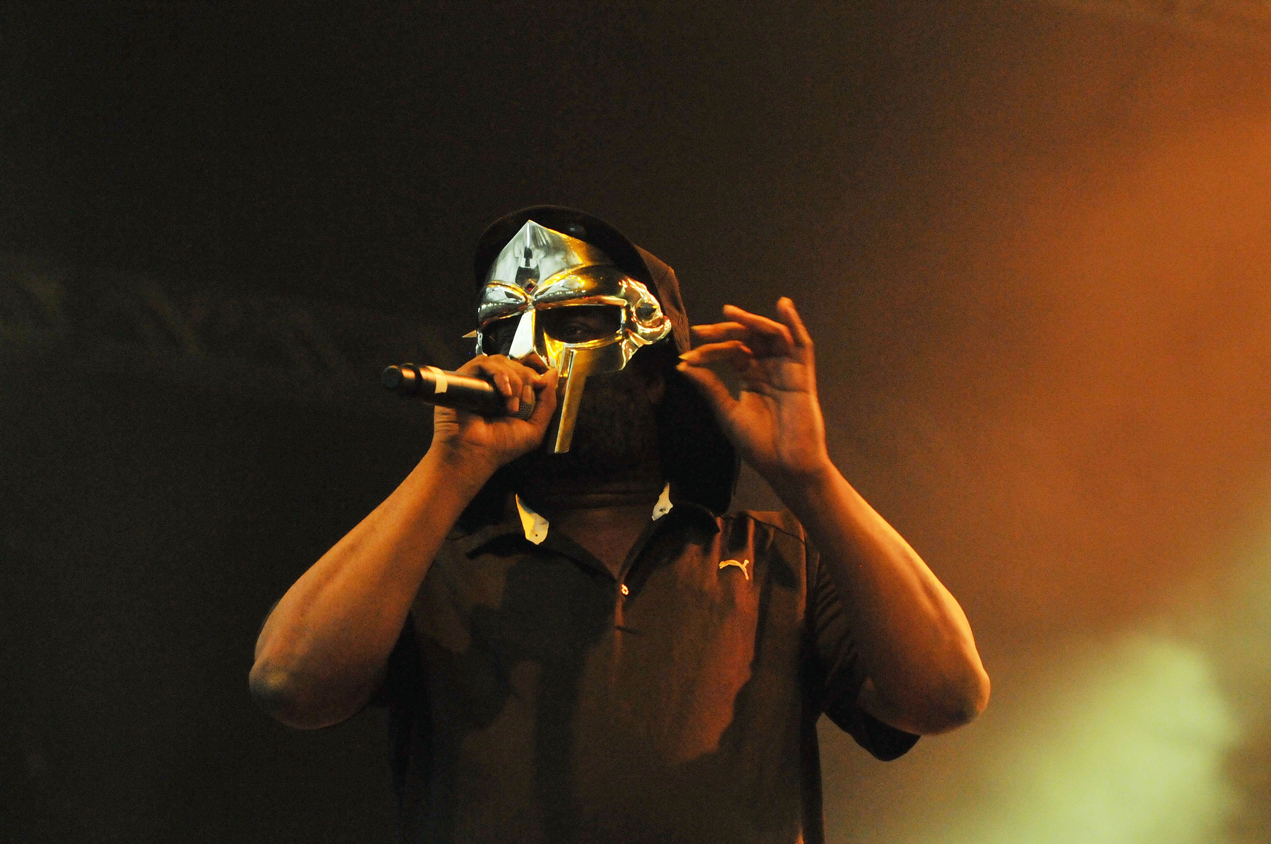 Rapper MF DOOM performing on stage, wearing his iconic metal mask, holding a microphone, with a dark background.