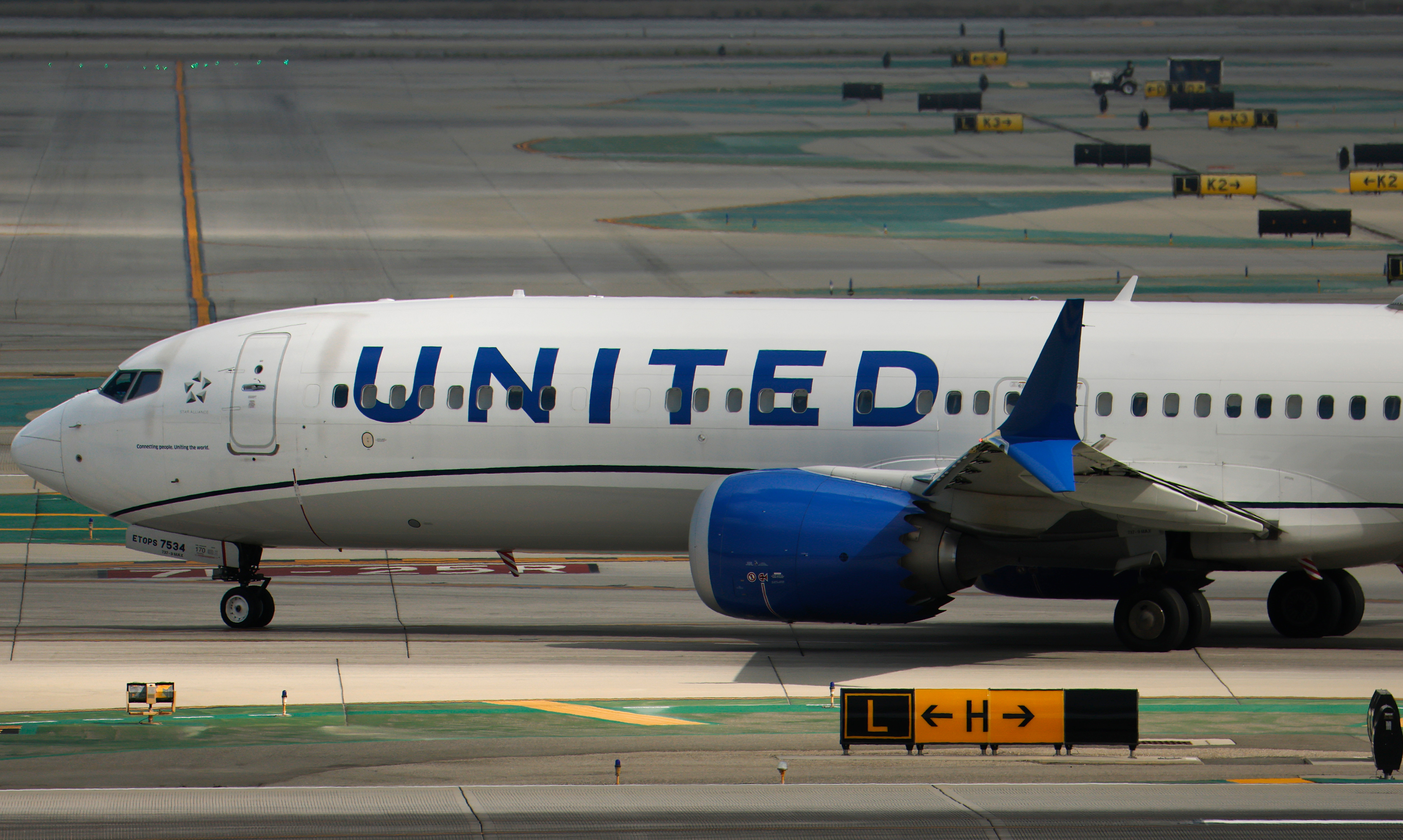 A United Airlines plane on a runway, with visible landing gear and blue engine. The airport tarmac and taxiway signs are in the background.