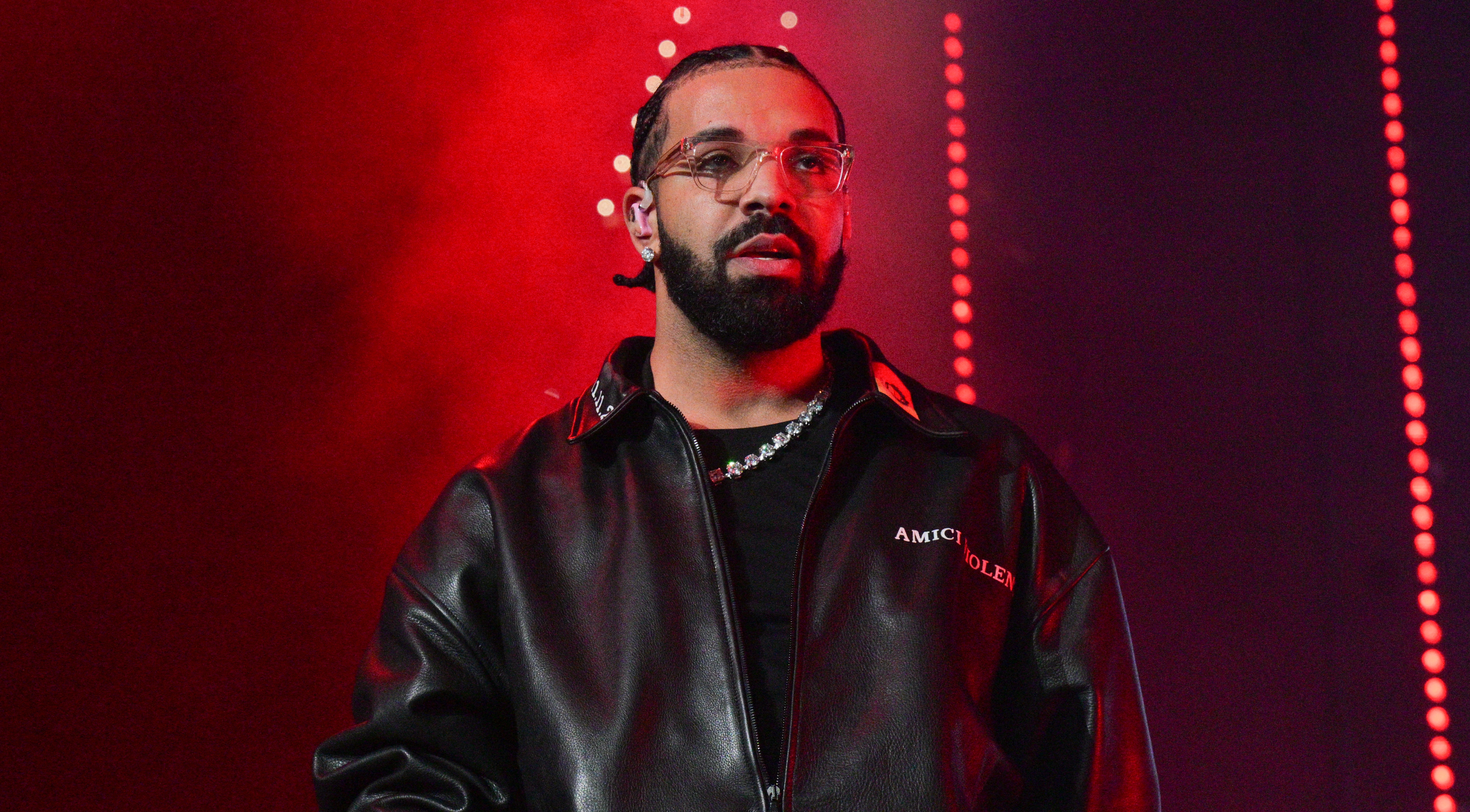 Drake with a beard and glasses performs on stage, wearing a black leather jacket, with red lighting in the background.