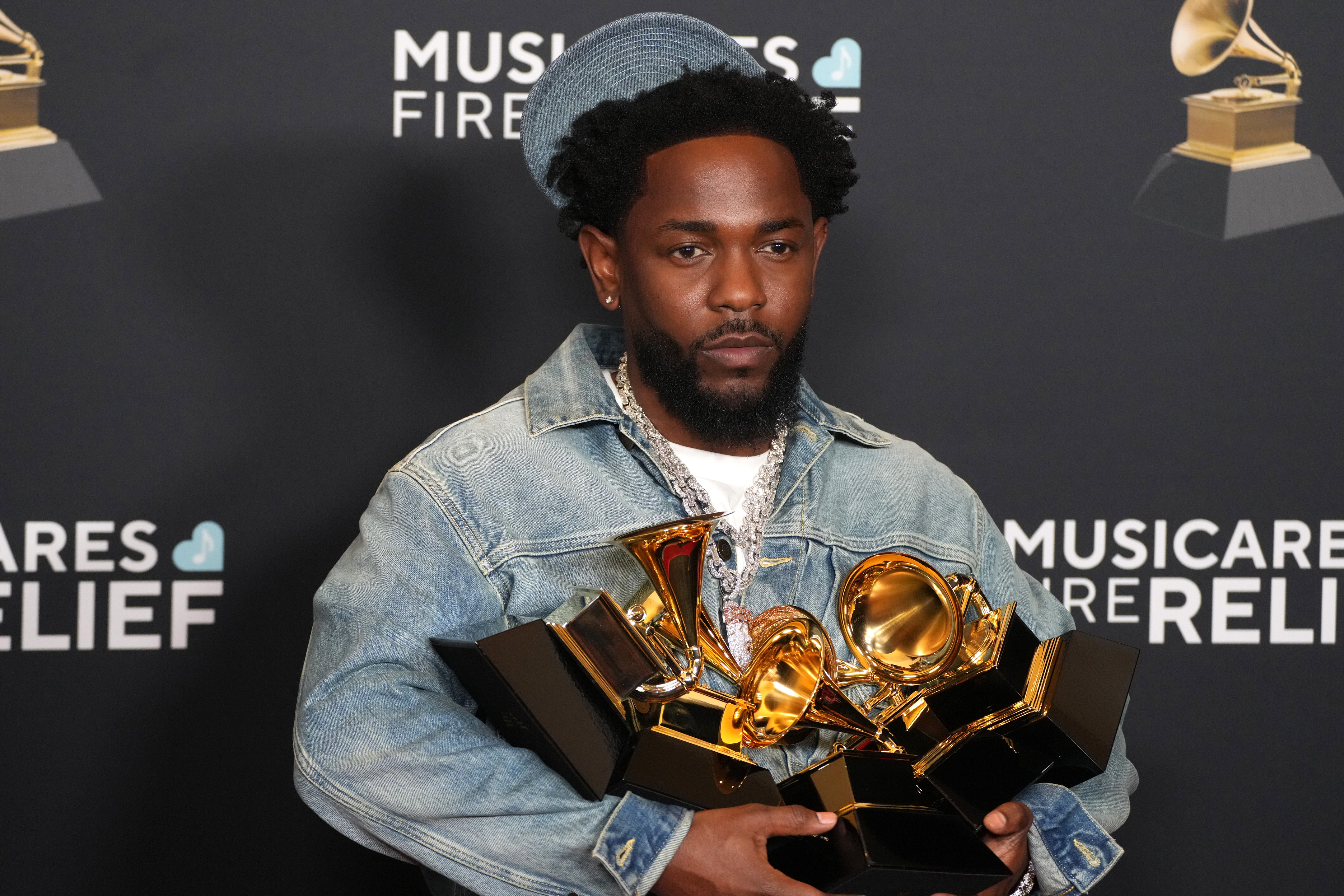 Kendrick Lamar holding multiple Grammy awards, wearing a denim jacket and hat, in front of a MusiCares Fire Relief backdrop.