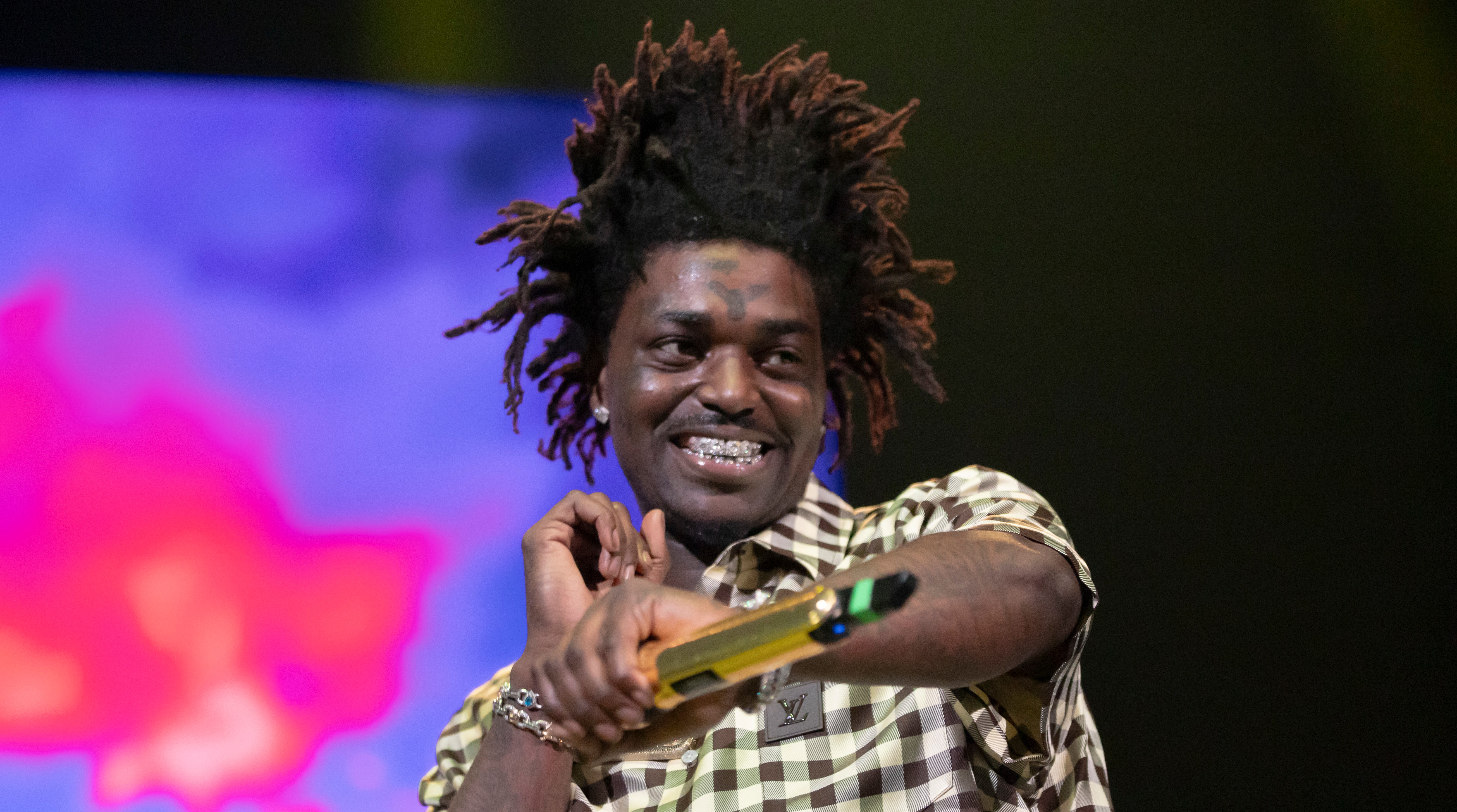 Kodak Black with distinctive hair and a checkered shirt holds a microphone on stage, smiling, with a colorful background.