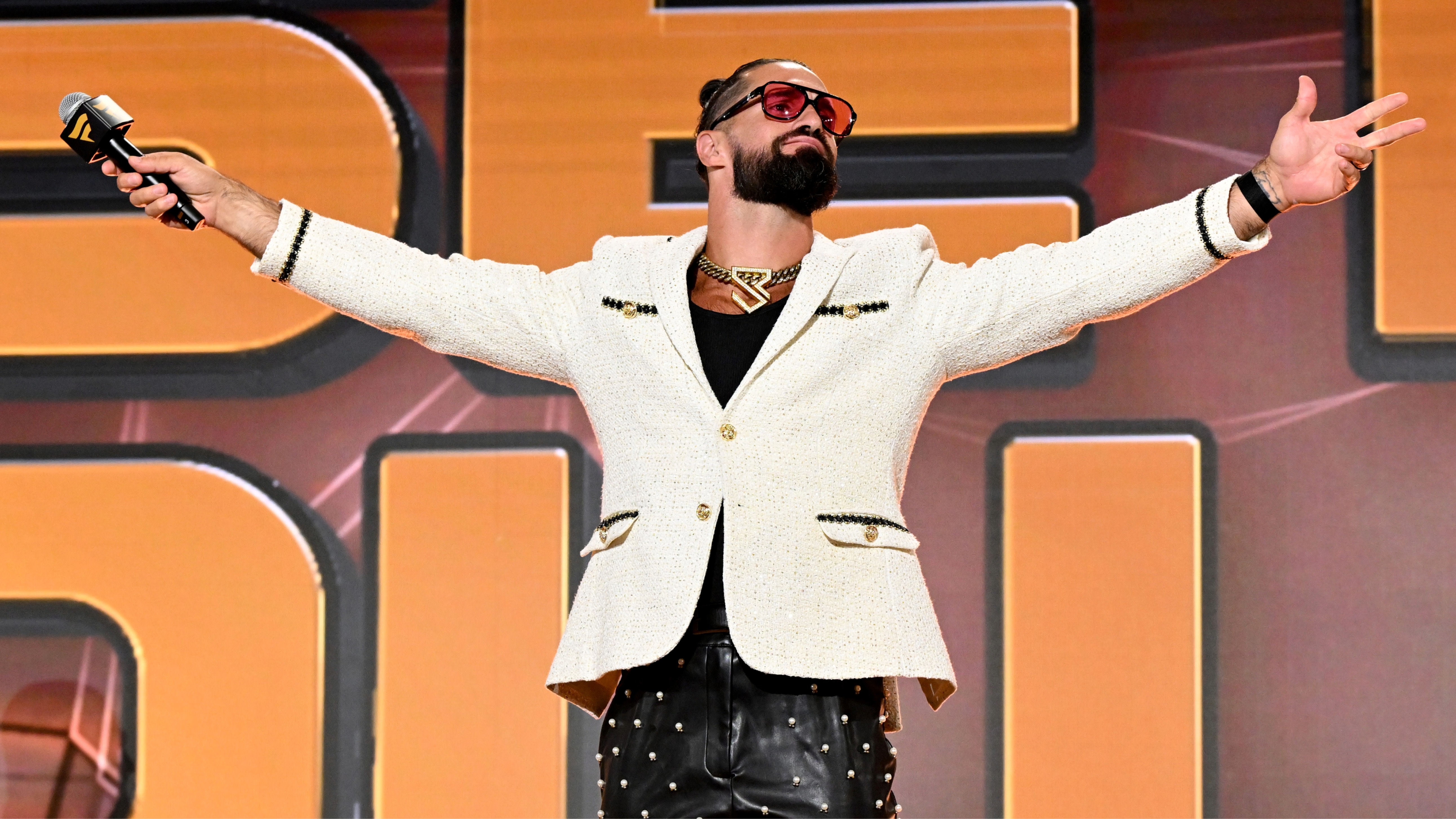 Seth Rollins in a white jacket and sunglasses, holding a microphone, stands with arms outstretched on stage.