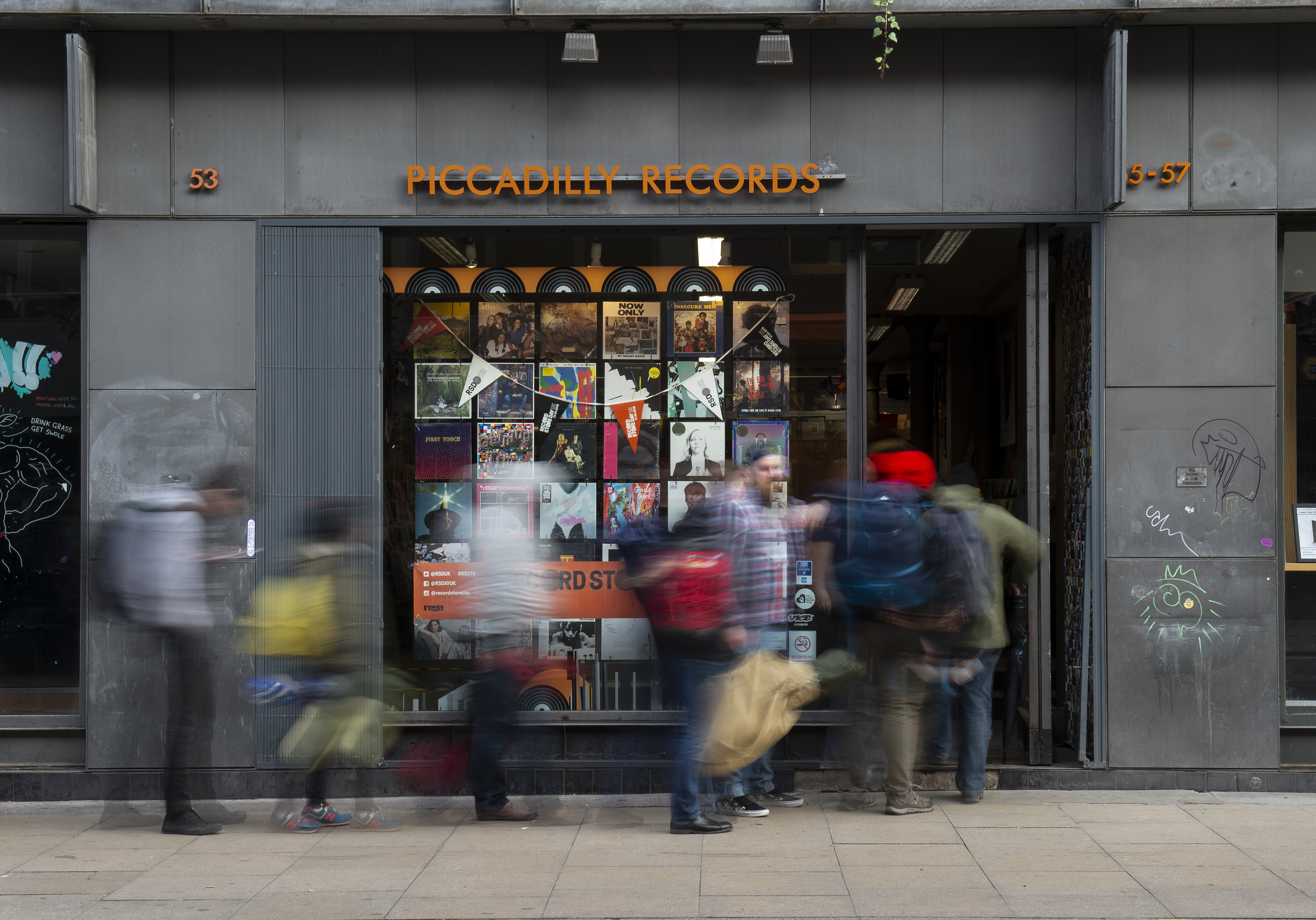 People walking past Piccadilly Records, a music store with a window display of album covers.