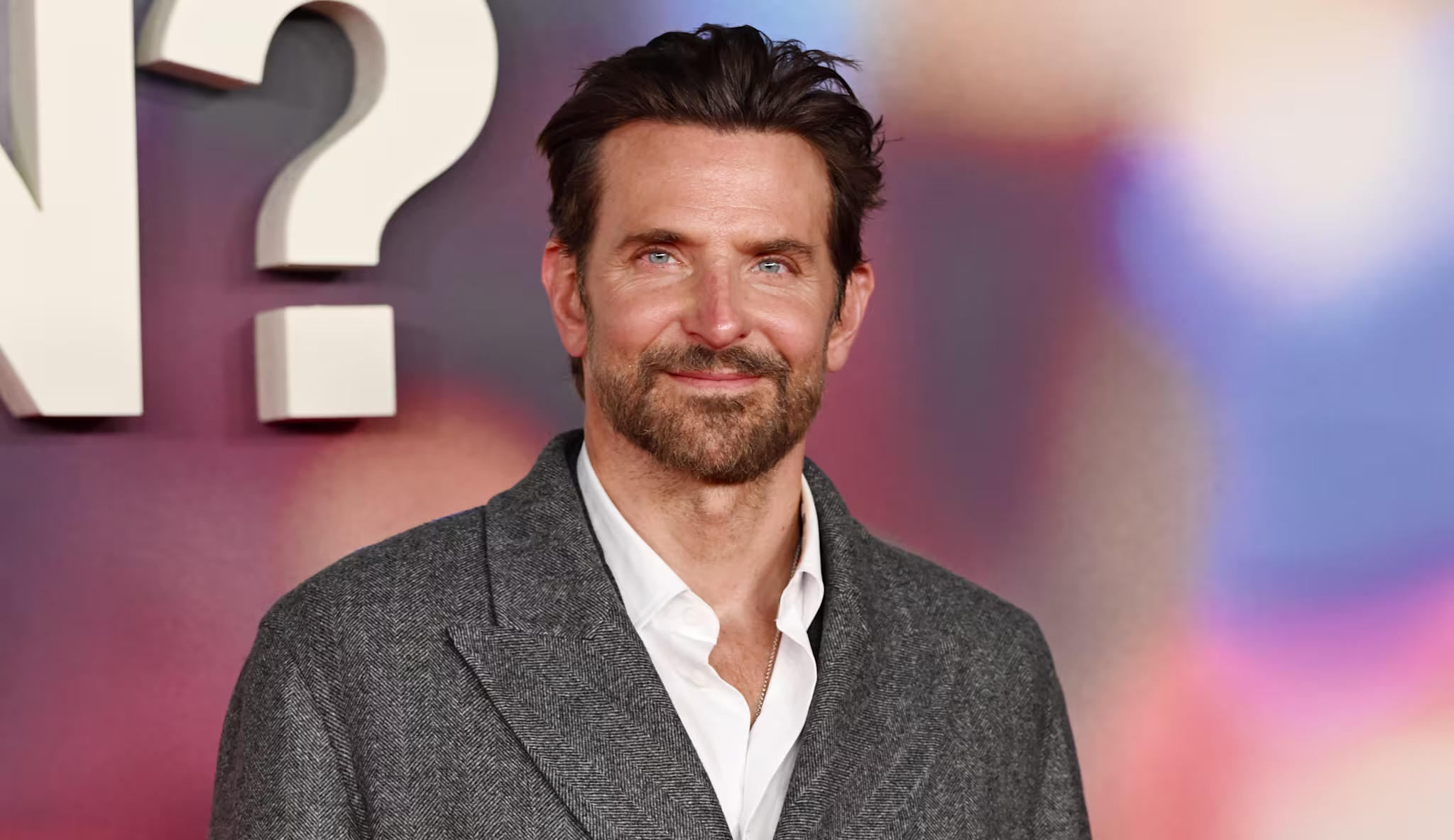 Bradley Cooper in a gray coat poses in front of a colorful blurred background.
