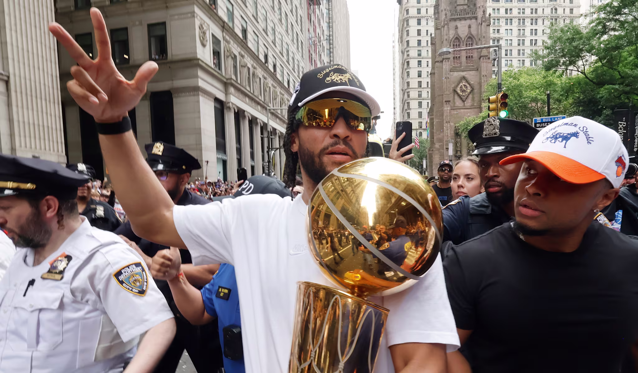 Jalen Brunson in sunglasses and a cap holds a basketball trophy during a parade, surrounded by people and police officers on a city street.