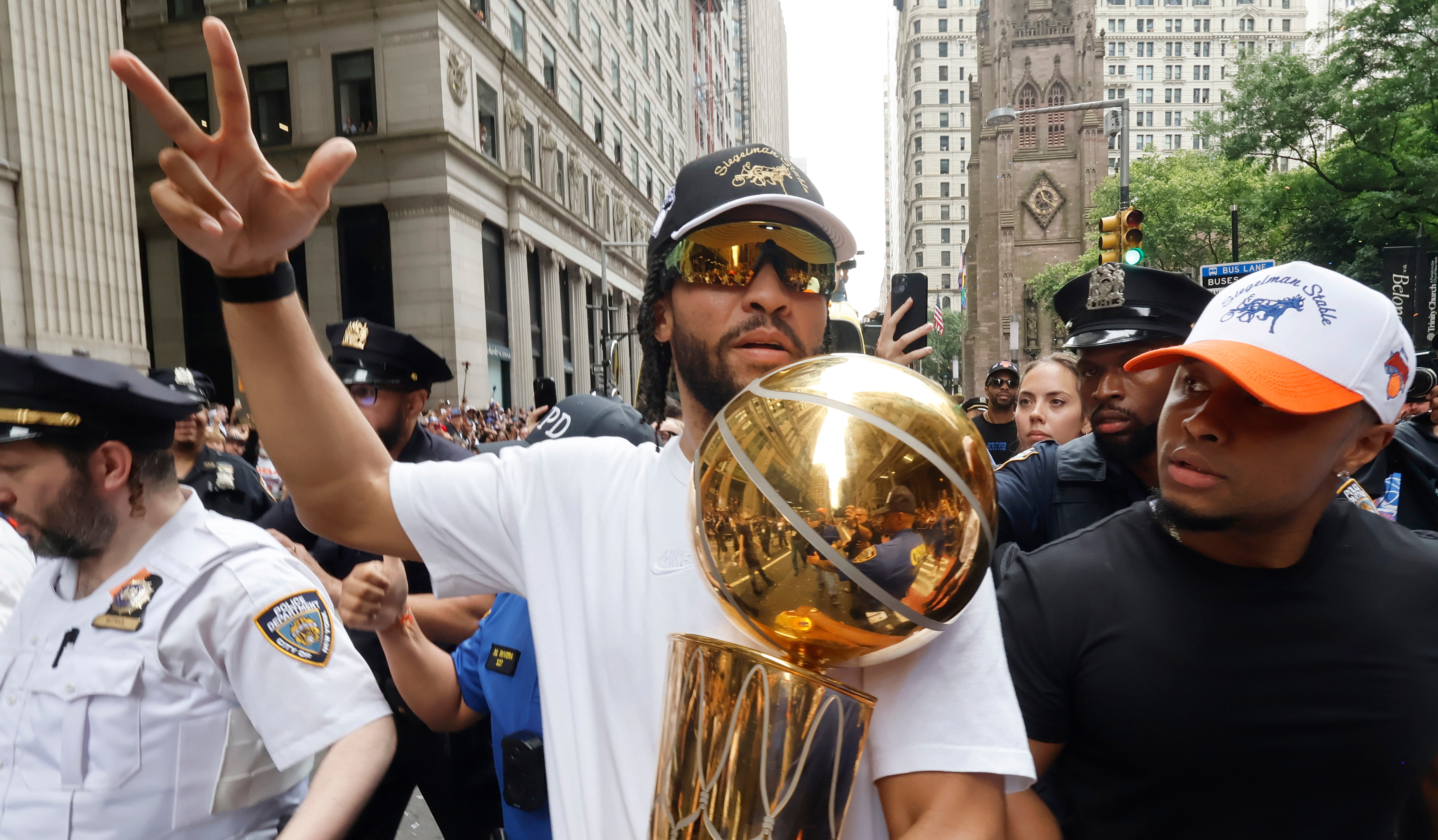 Jalen Brunson holding a championship trophy in a parade, wearing sunglasses and a cap, surrounded by people and police officers in a city setting.