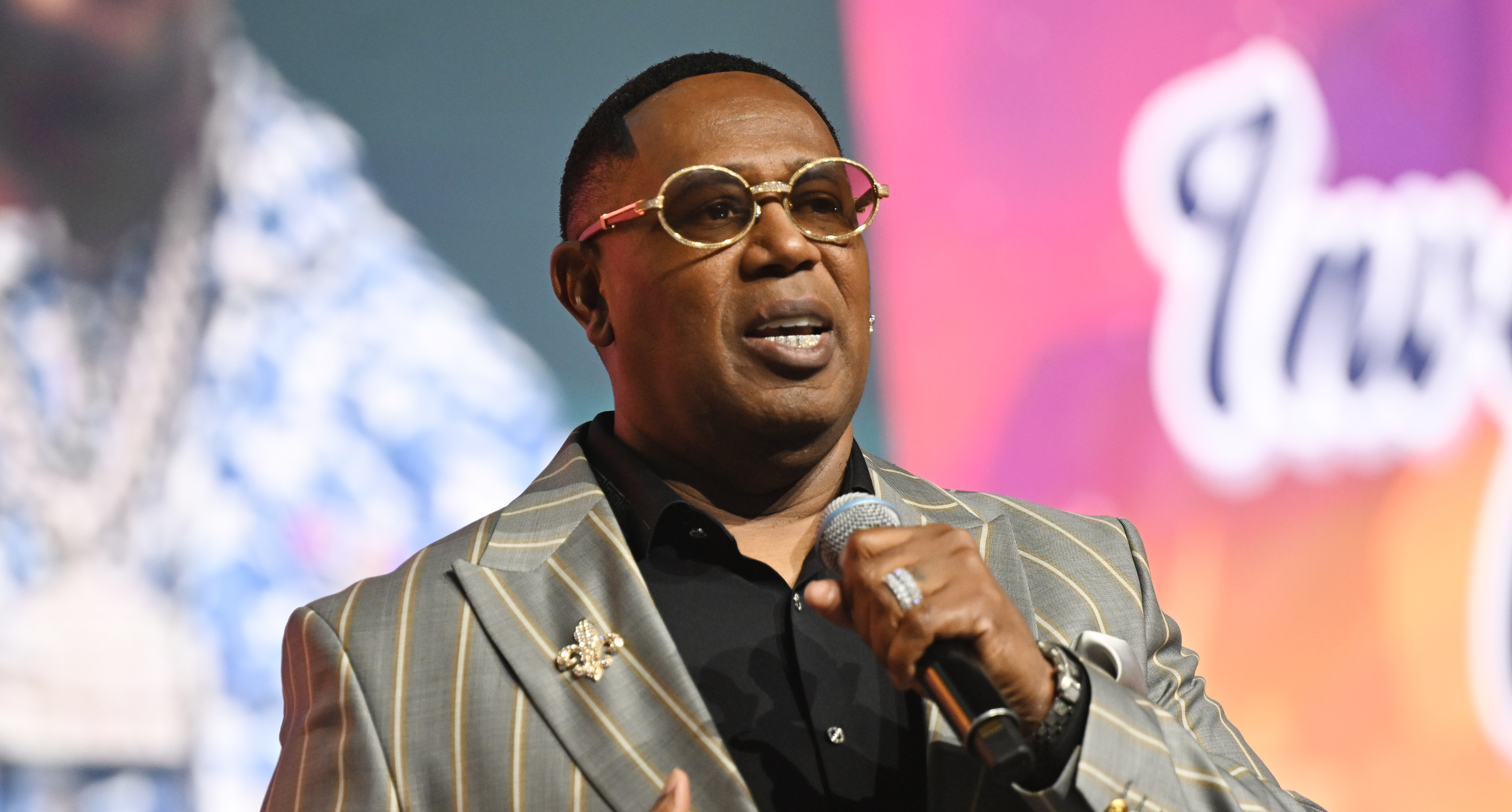 Master P speaking on stage, wearing a striped suit and sunglasses, holding a microphone.