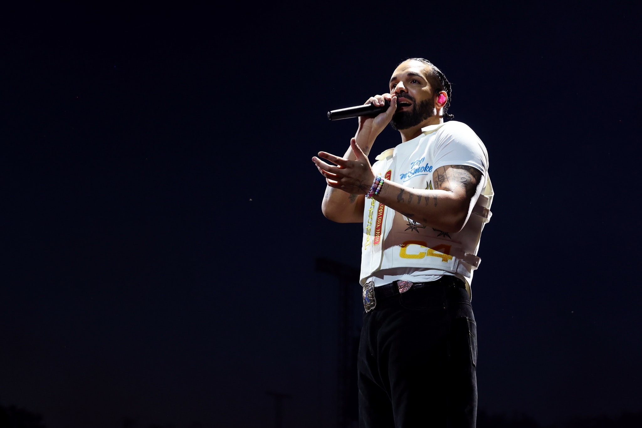 A man with a beard and braided hair performs on stage, holding a microphone, wearing a white t-shirt and black pants, against a dark background.
