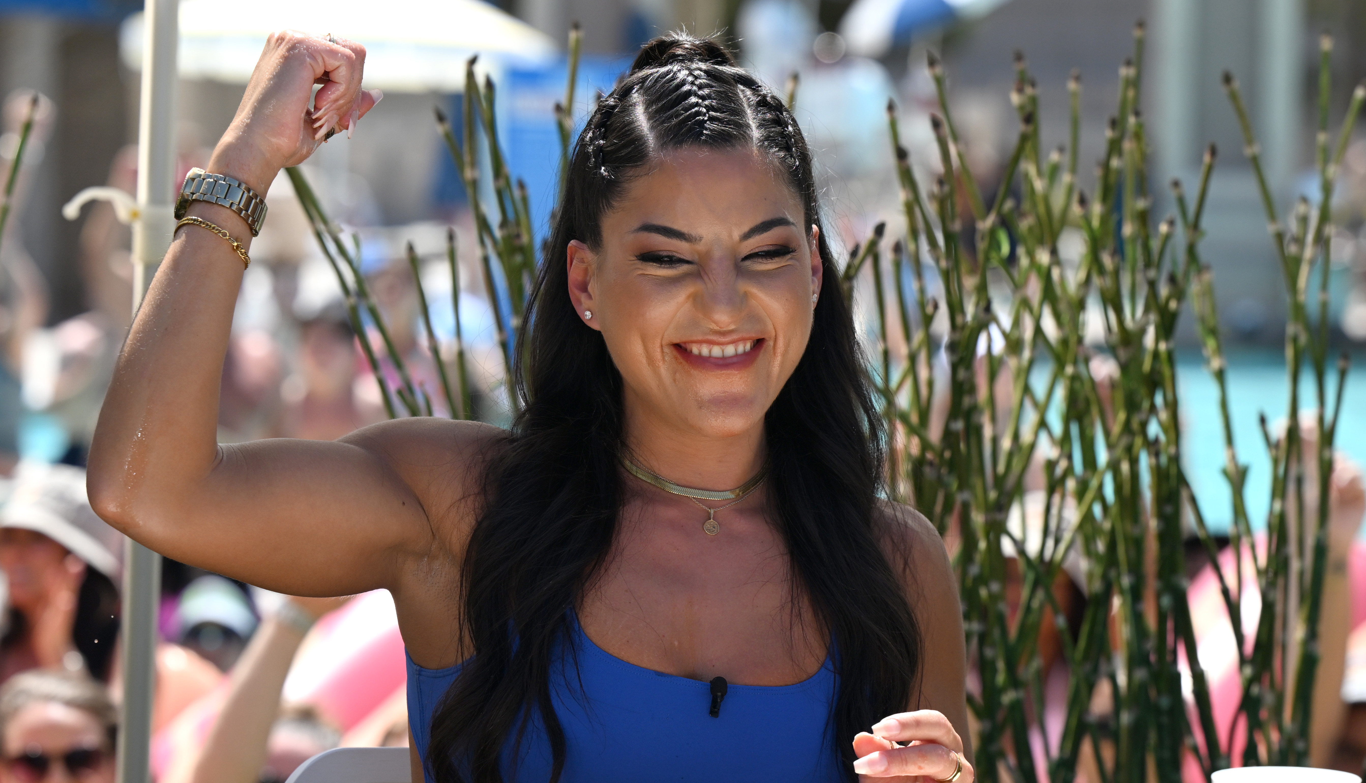 Leah Shutkever in a blue outfit, smiling and raising her fist, sits at a table with a tray, surrounded by a crowd and greenery.
