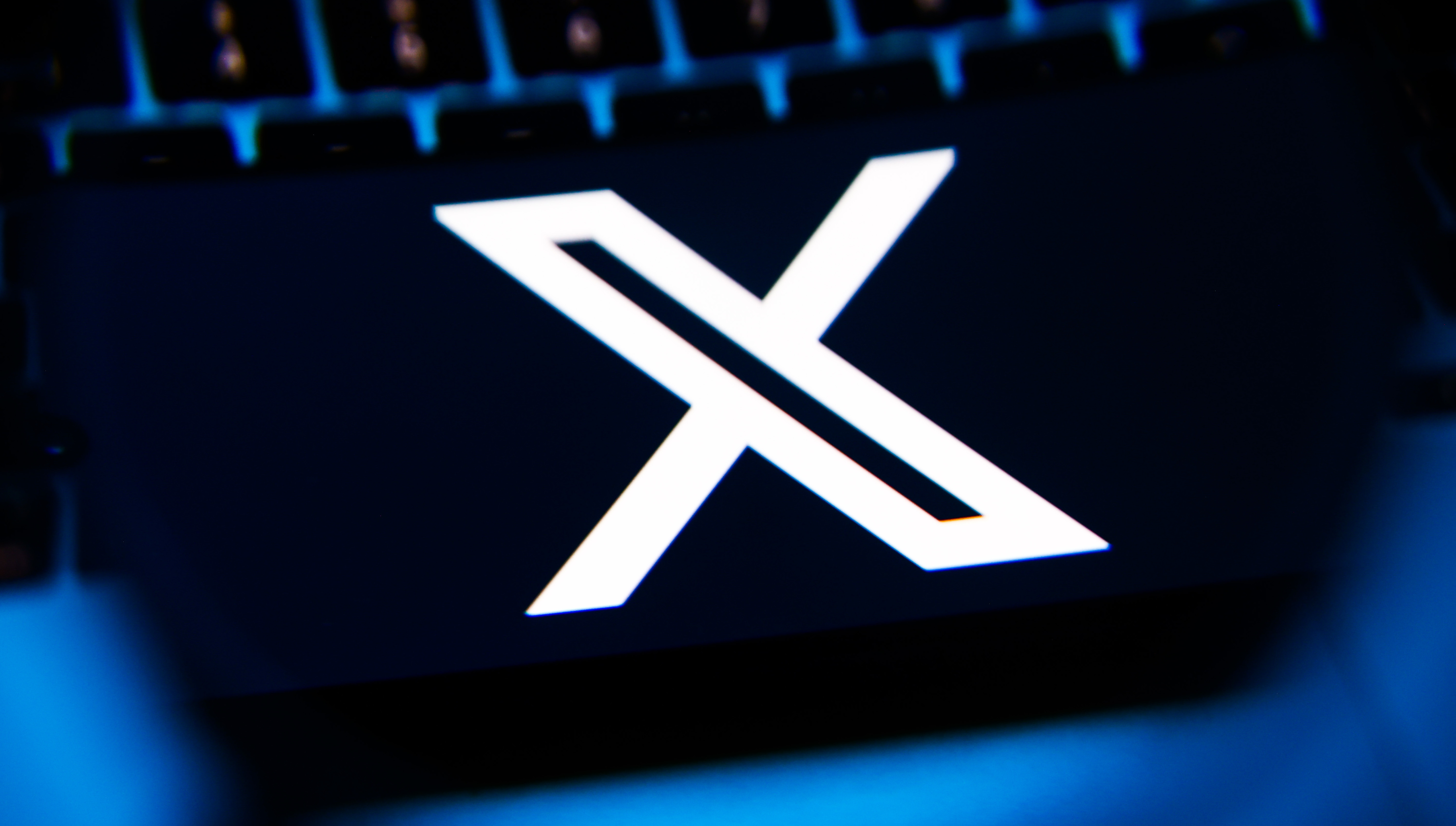A white "X" logo on a dark background, with a blurred keyboard in the background, illuminated by blue light.