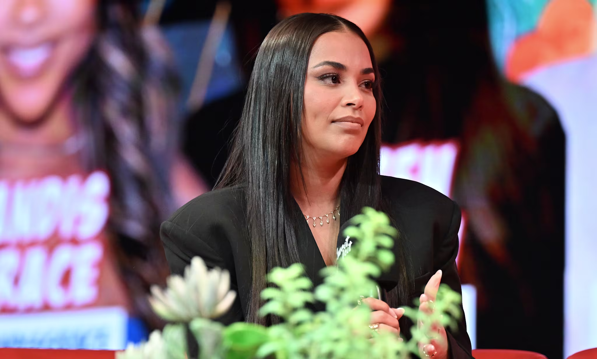 Lauren London with long dark hair sits on a stage, wearing a black blazer. 