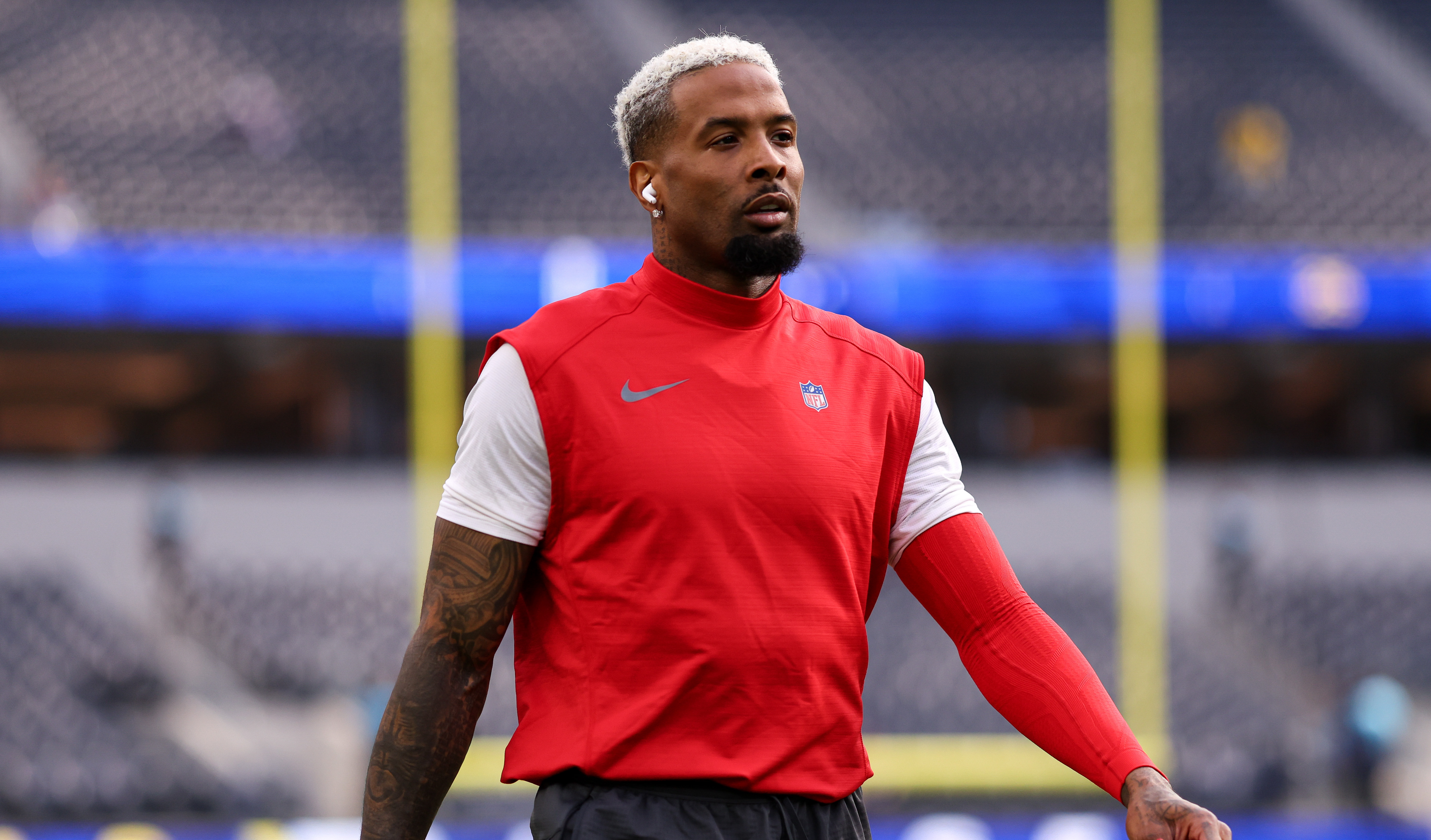 Odell Beckham Jr. with bleached hair, wearing a red sleeveless top and shorts, walks on a field holding red gloves. Stadium background.