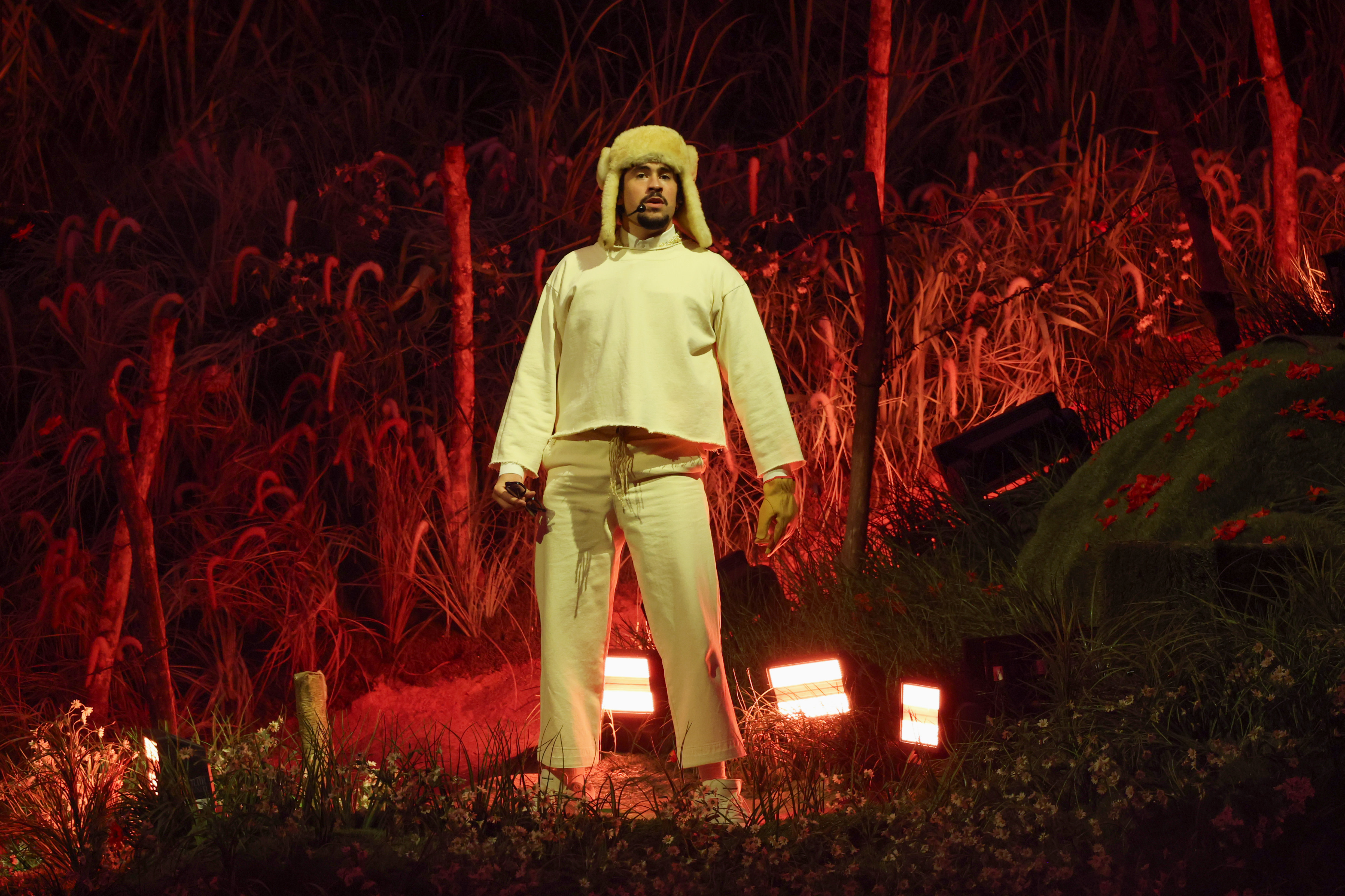 Bad Bunny in white clothing and a fur hat stands on a stage with a forest backdrop and red lighting.