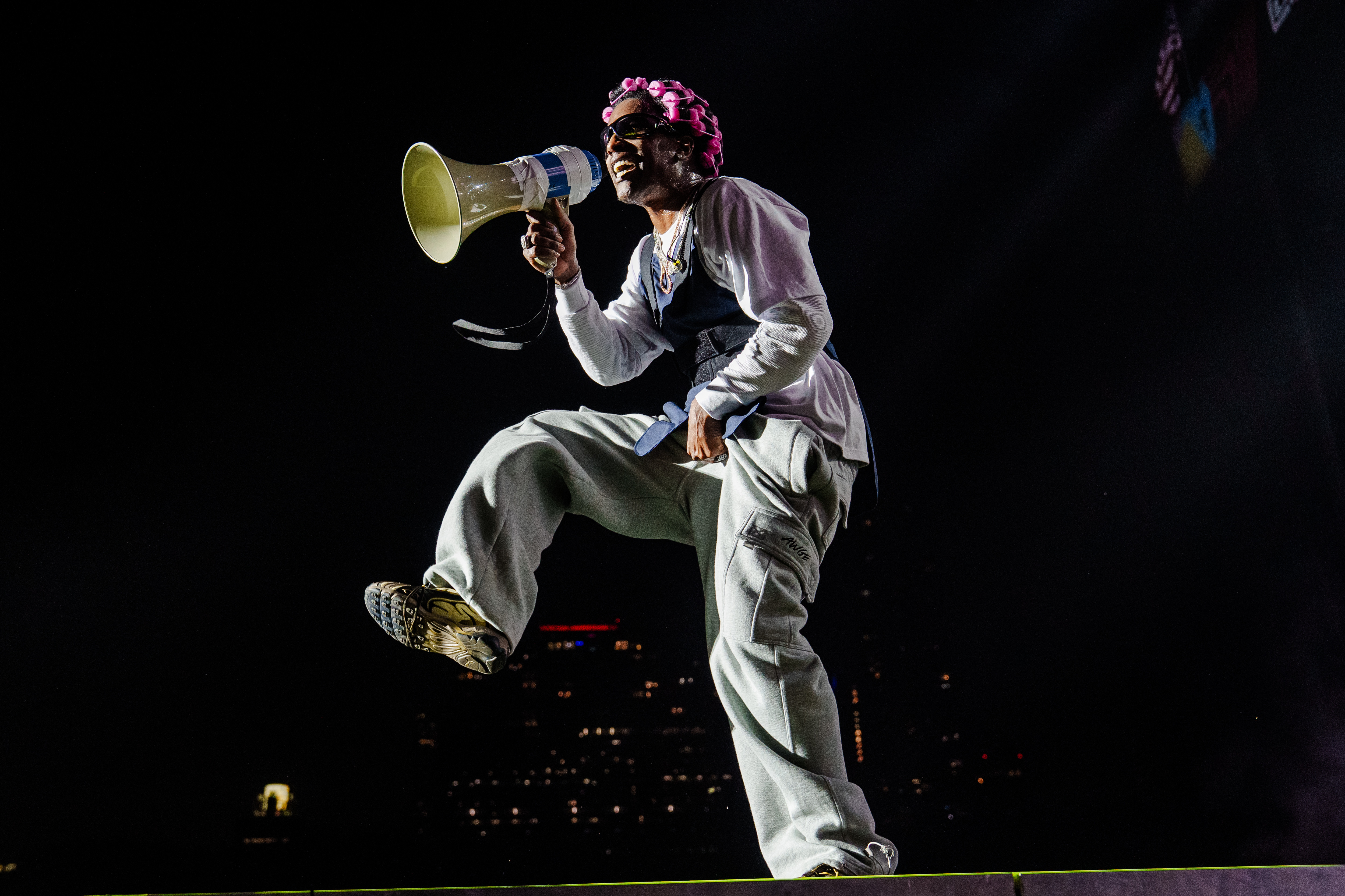 ASAP Rocky with a megaphone, wearing a pink headpiece and casual outfit, energetically poses on stage against a dark cityscape. This performance was in support of his new album, 'Don't Be Dumb.'