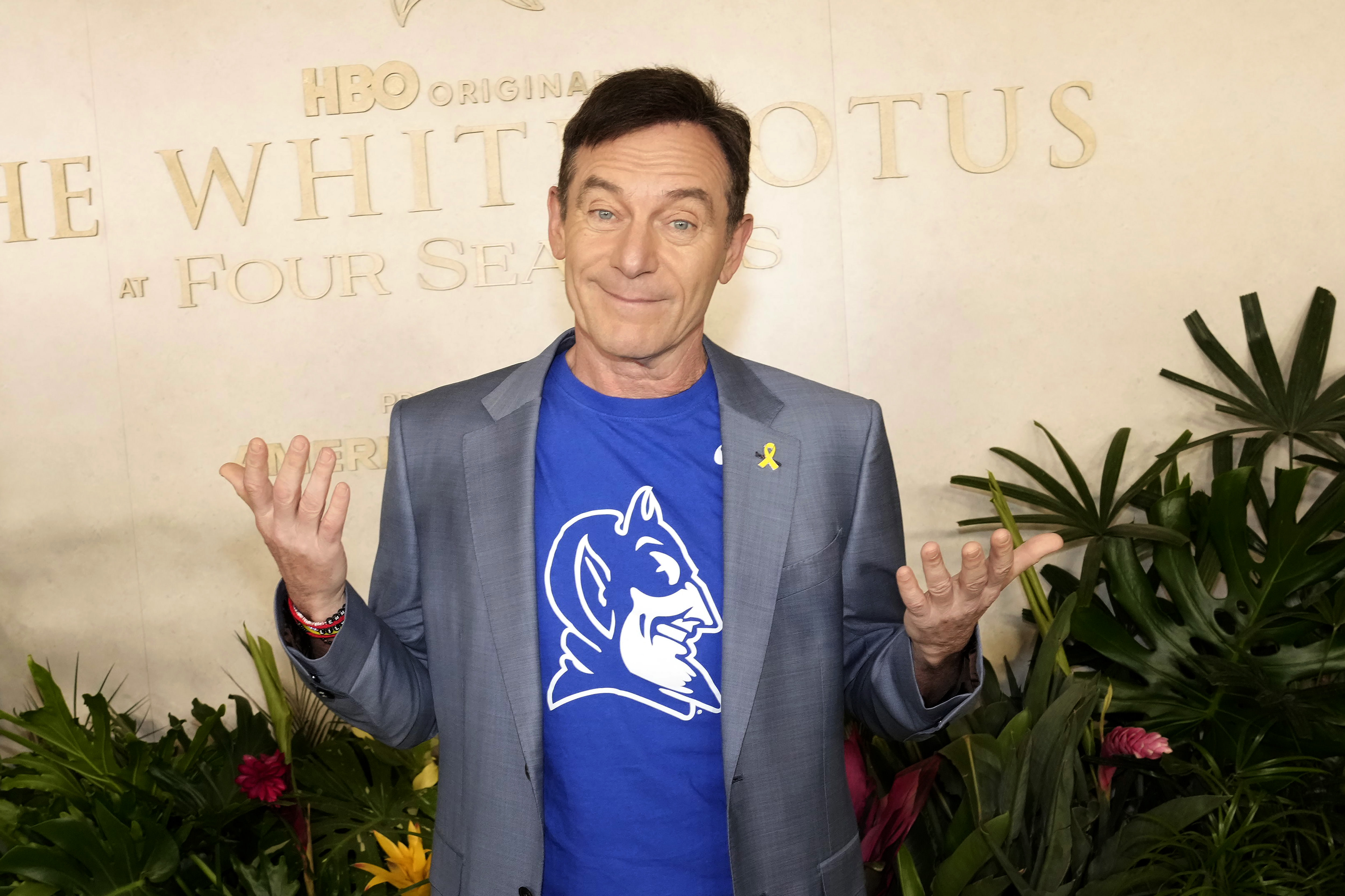 Jason Isaacs in a gray blazer and blue shirt standing in front of a "The White Lotus" backdrop, surrounded by plants.