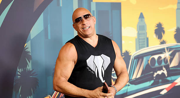 Vin Diesel, wearing sunglasses and a sleeveless shirt, smiles in front of a colorful backdrop with cars and palm trees.