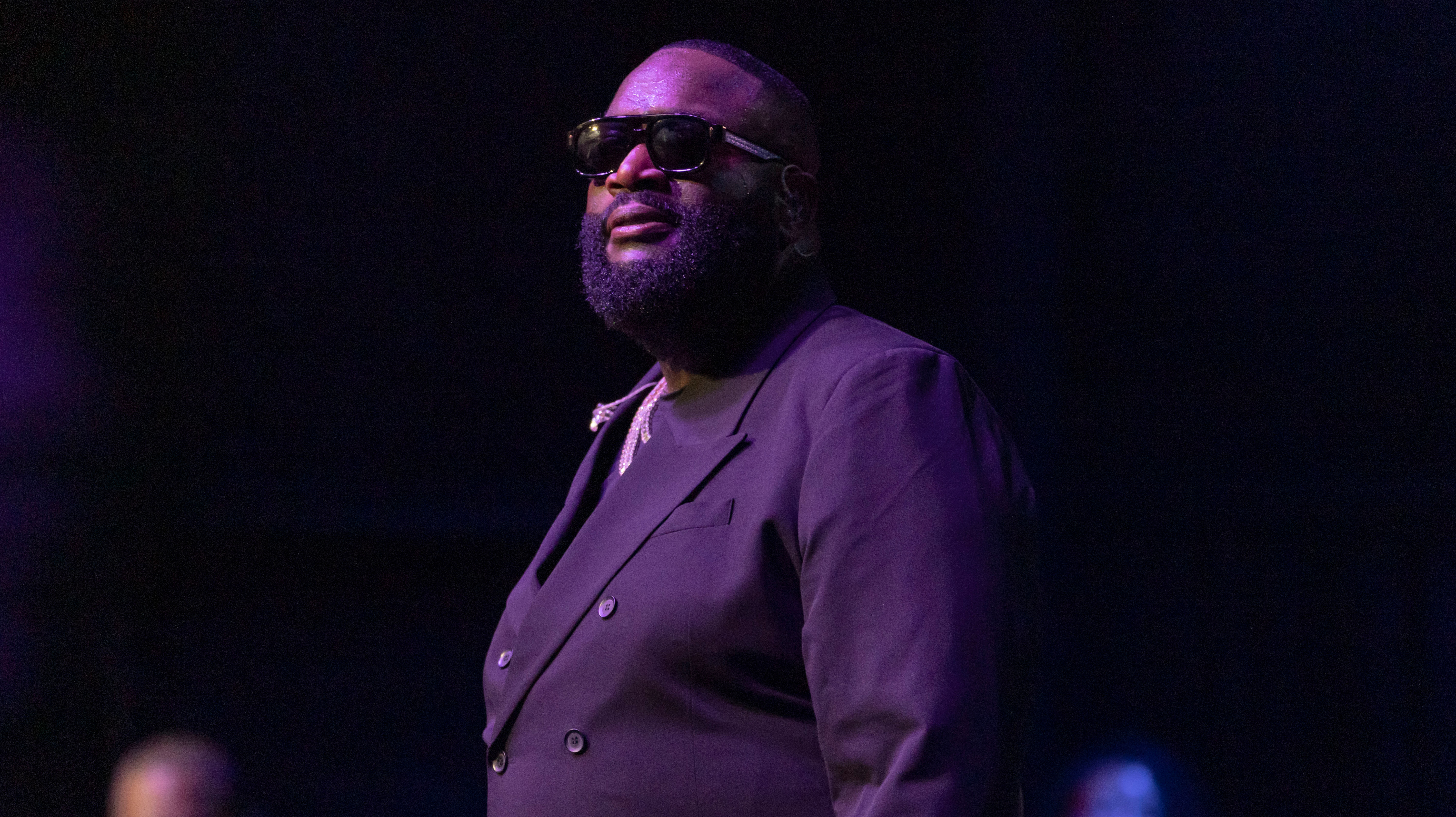 Rick Ross in a dark suit and sunglasses stands on stage with a confident expression, under purple lighting.