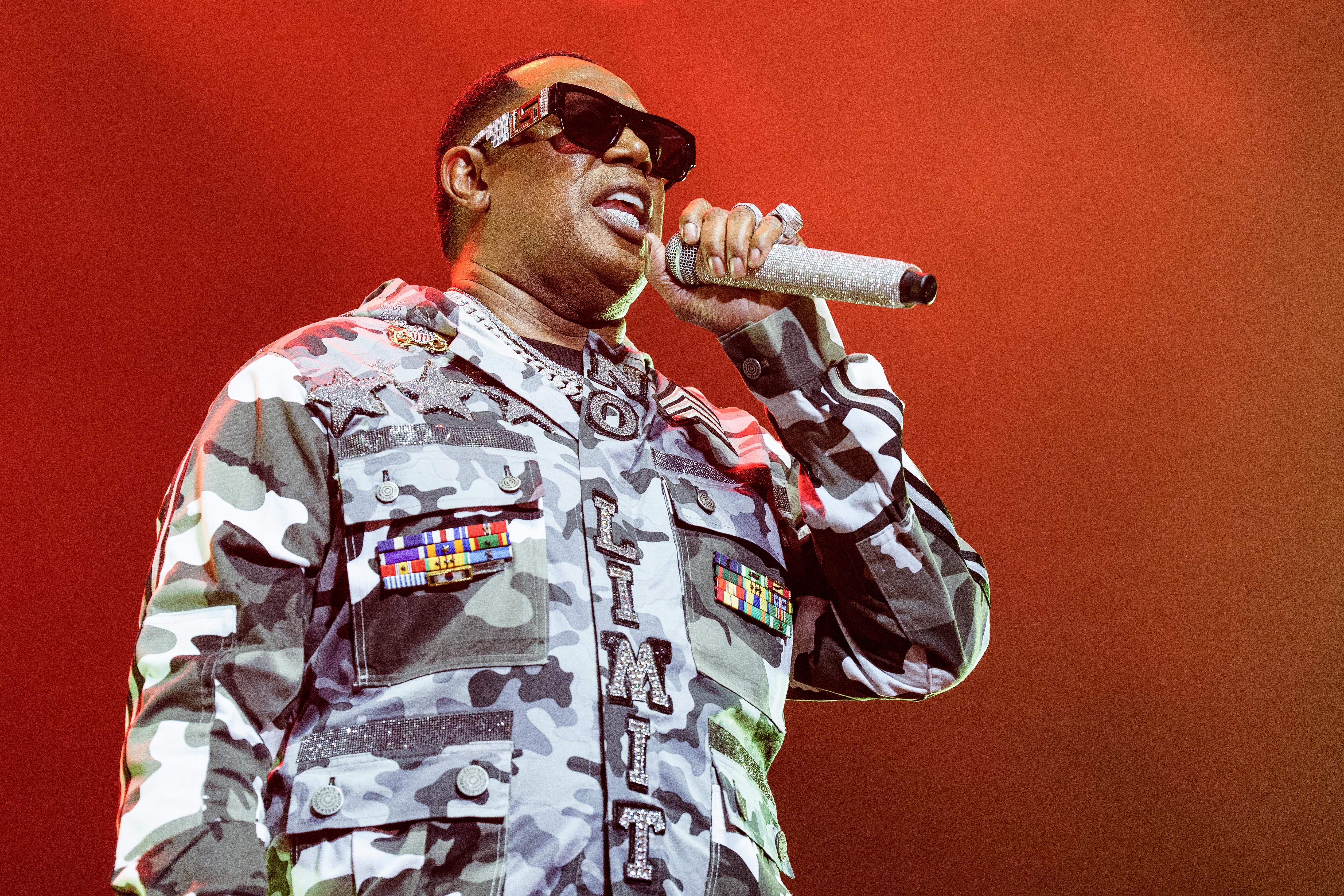 A performer in a camo jacket with "No Limit" performs on stage, holding a sparkly microphone, against a red background.