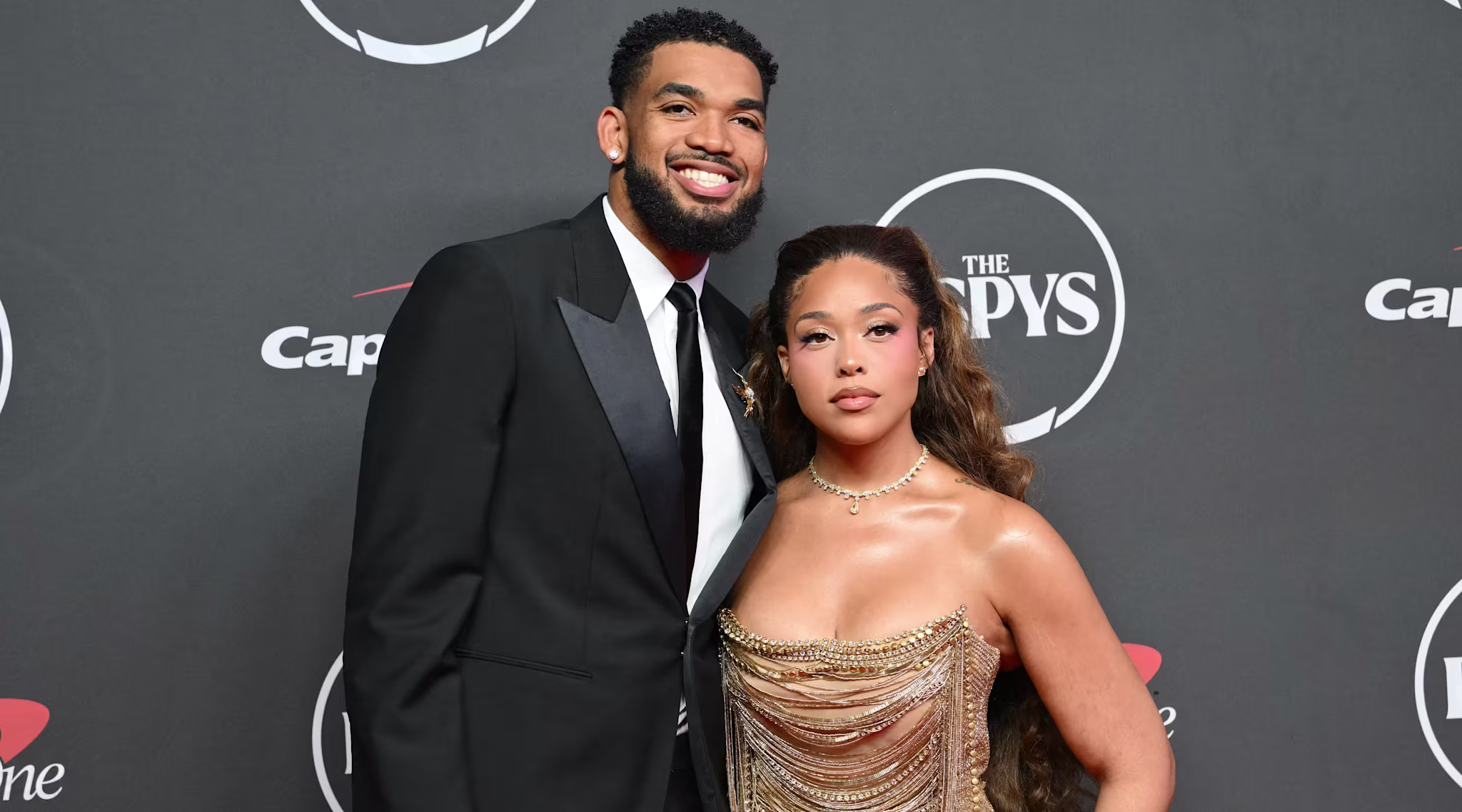 Karl-Anthony Towns in a black tuxedo and Jordyn Woods in a gold dress pose together on the red carpet at the ESPY Awards.