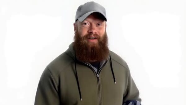 A bearded man wearing a gray cap and green hoodie stands against a white background.