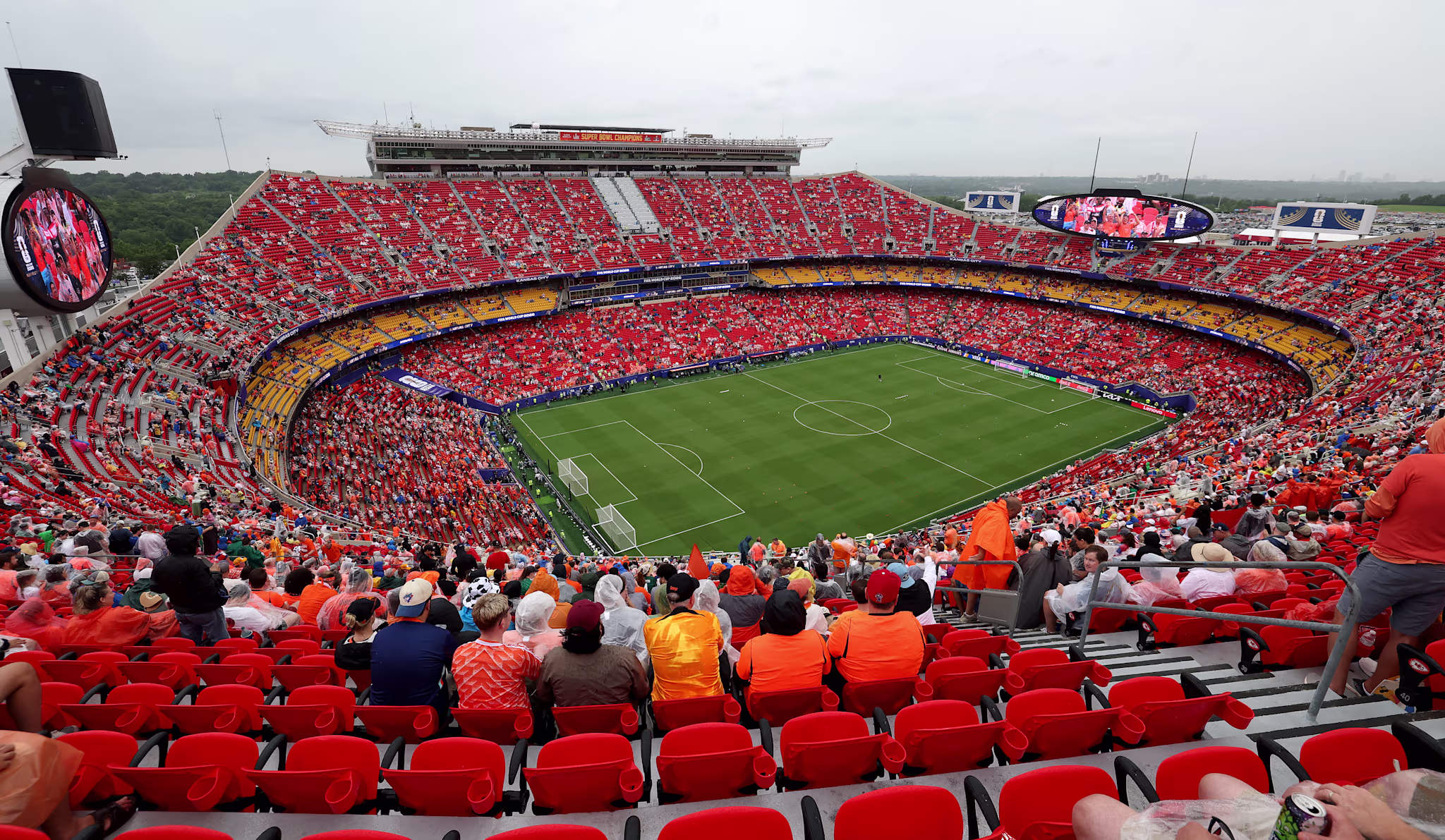 A packed soccer stadium with fans in orange and red seats, overlooking the field. Overcast sky visible in the background.