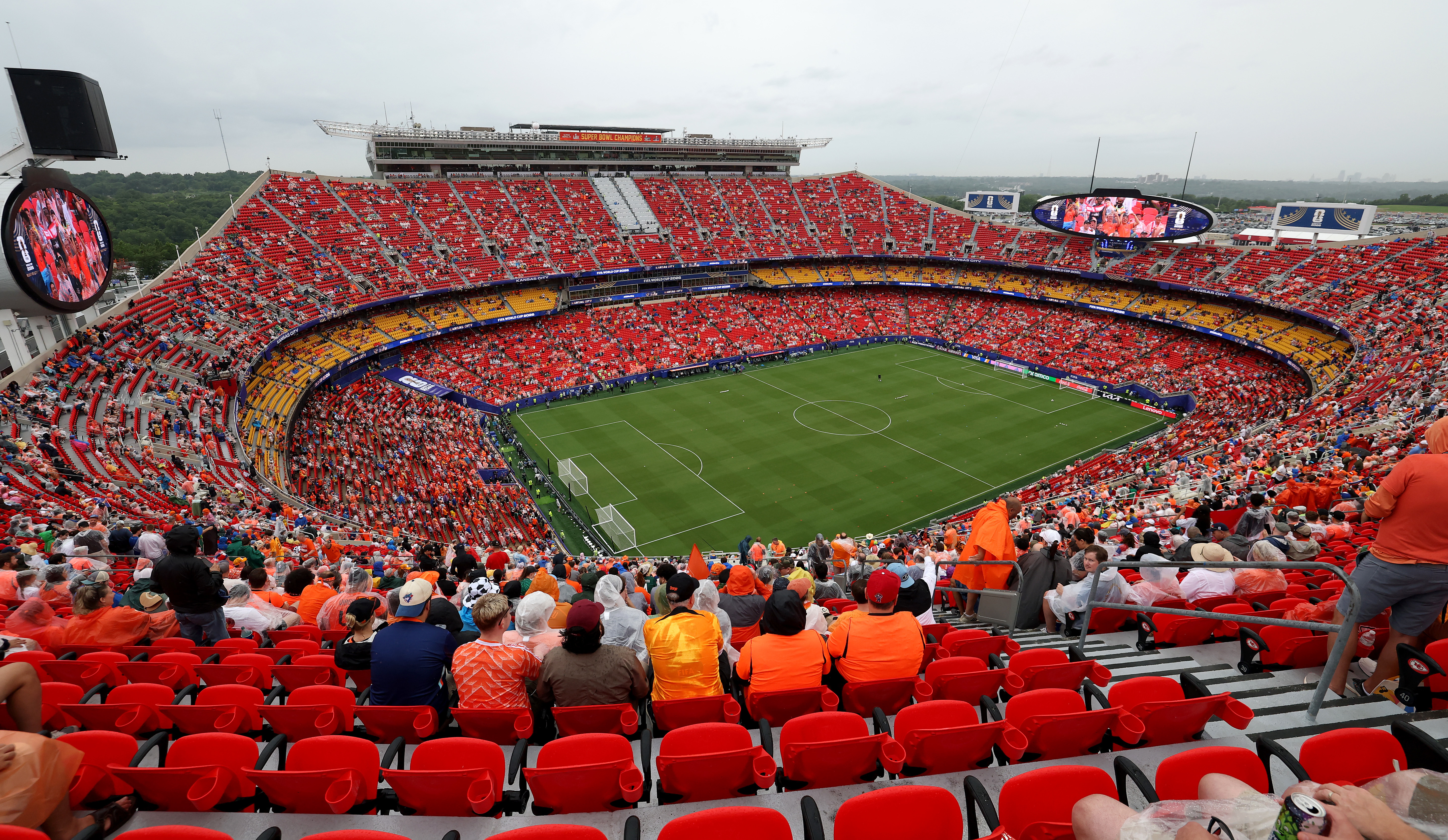 A packed soccer stadium with fans in orange and red seats, overlooking the field. Overcast sky visible in the background.