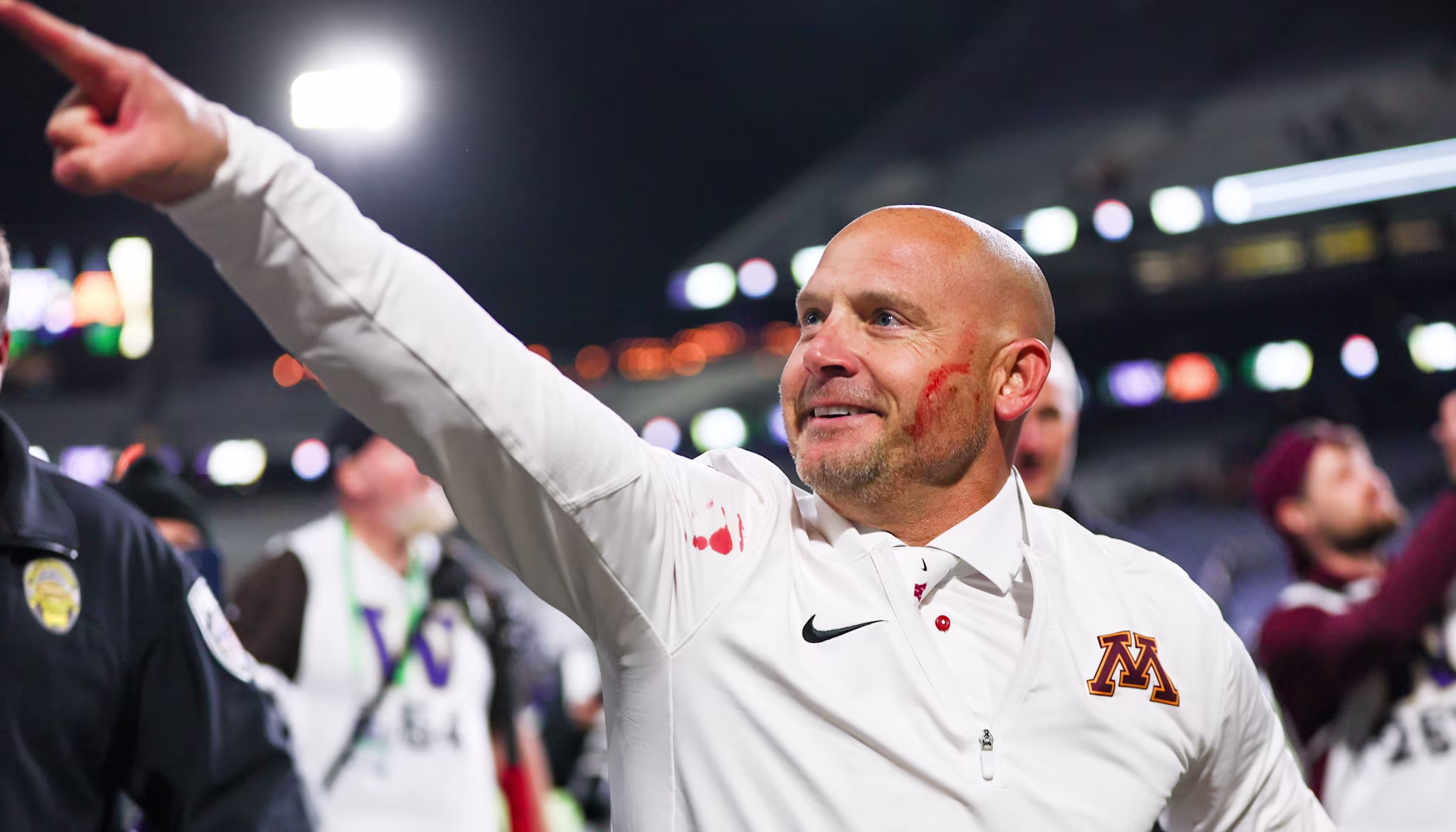 P.J. Fleck in a white Minnesota sports jacket, with a joyful expression, points forward in a stadium setting.