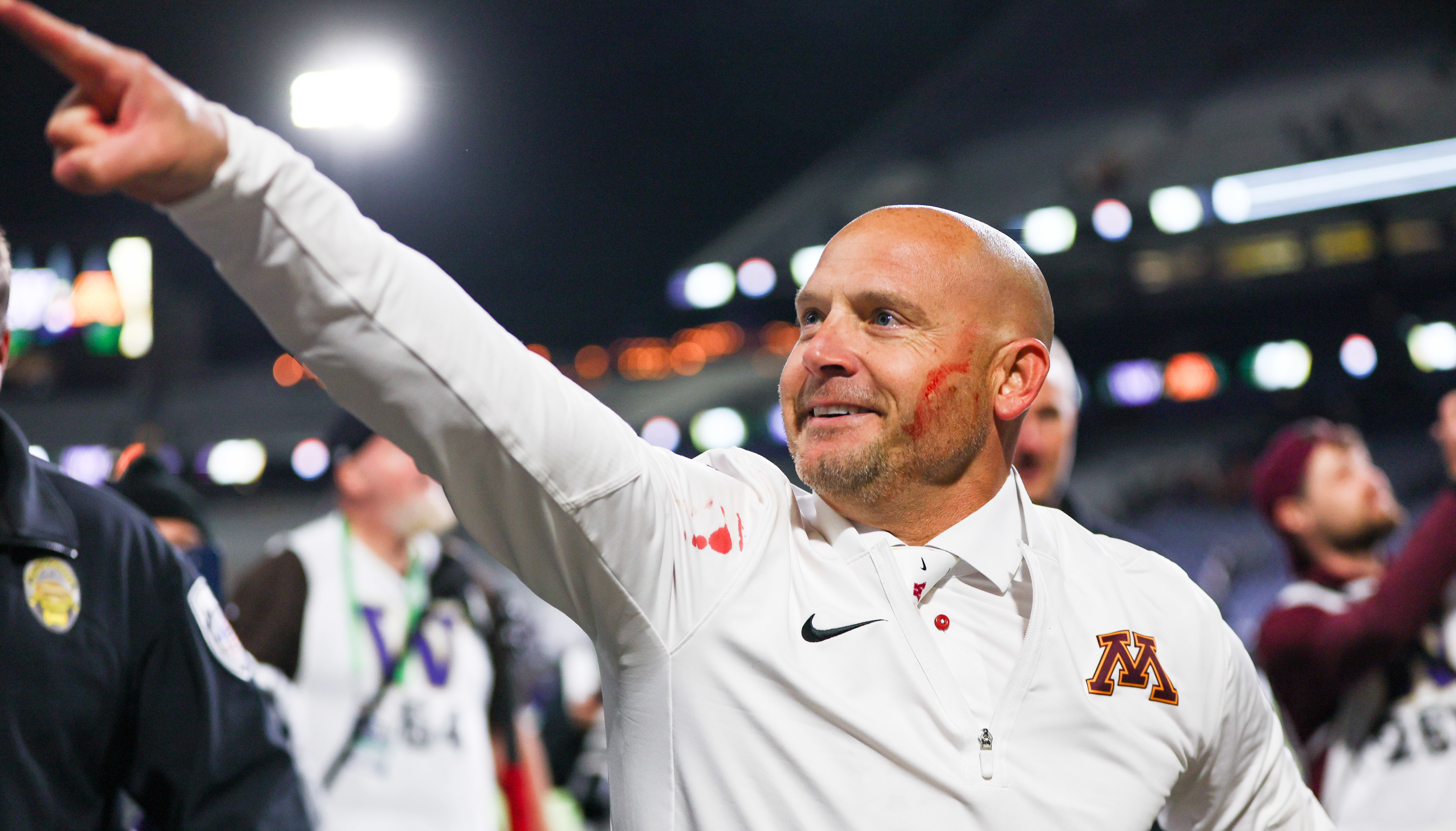 P.J. Fleck in a white Minnesota Golden Gophers jacket gestures enthusiastically in a stadium setting, with people and lights in the background.