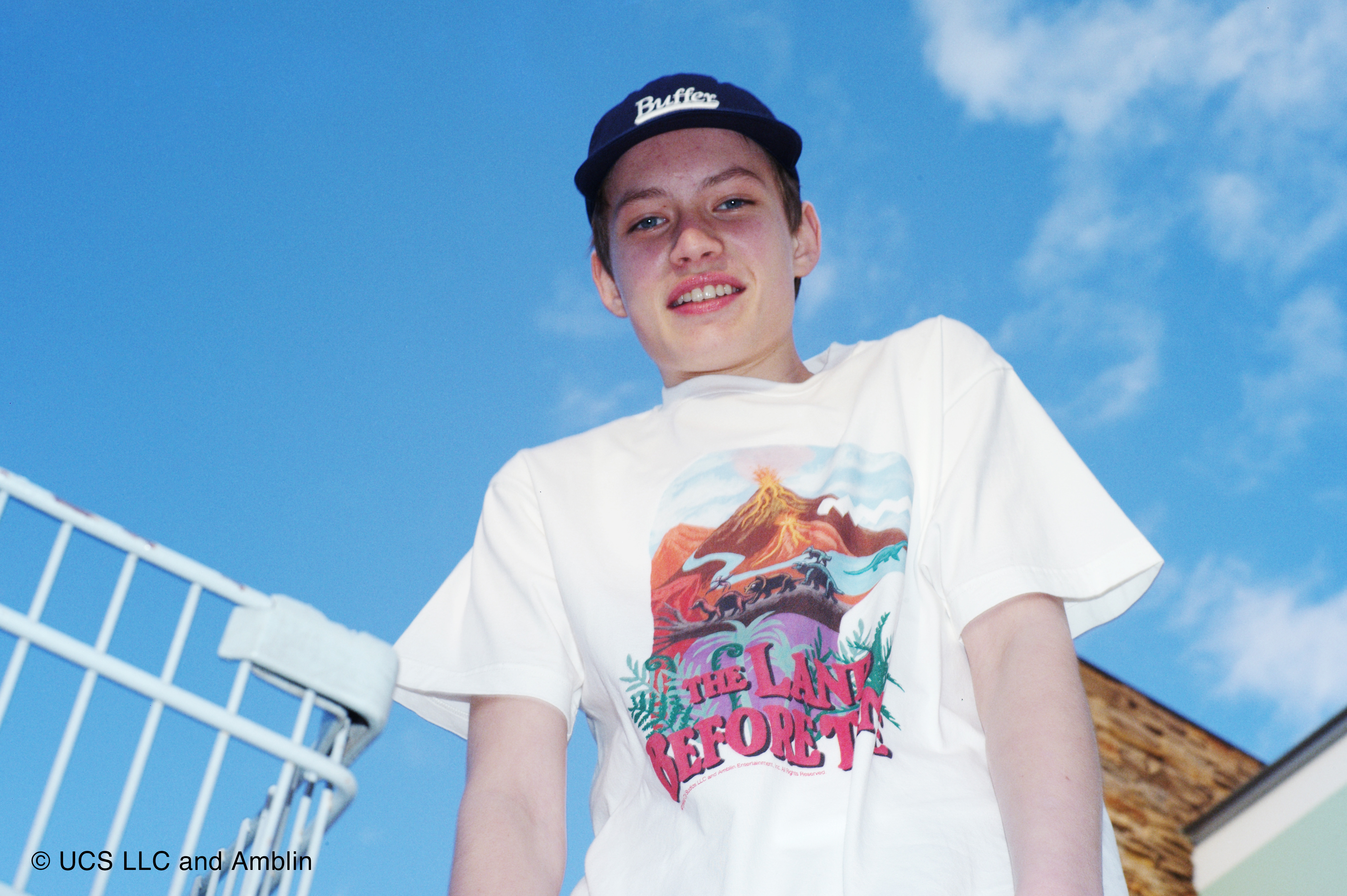 A young person wearing a "The Land Before Time" T-shirt and a blue cap smiles at the camera, with a shopping cart and blue sky in the background.