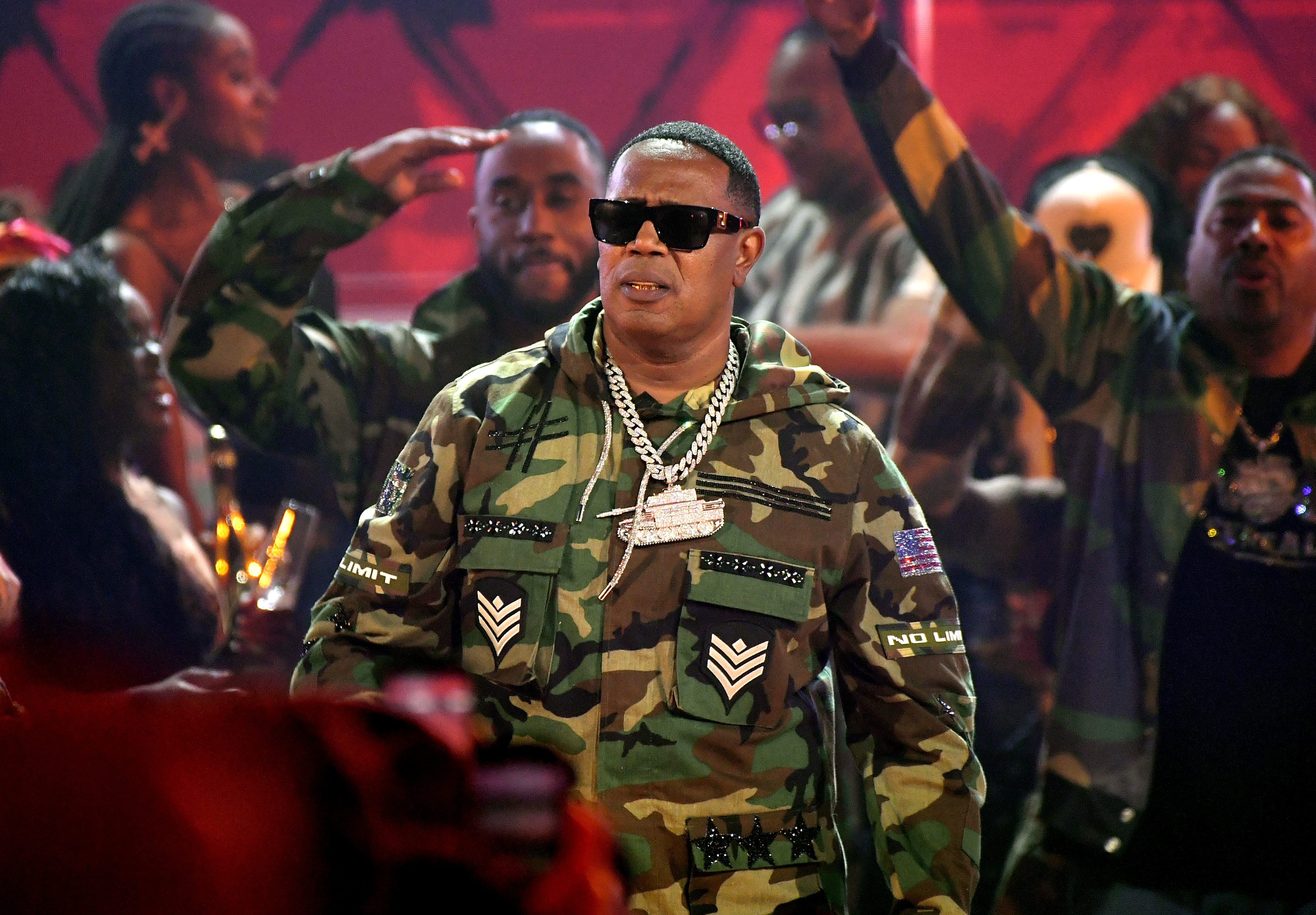 Master P in camo outfit, wearing sunglasses and a chain, surrounded by people in similar attire on stage.