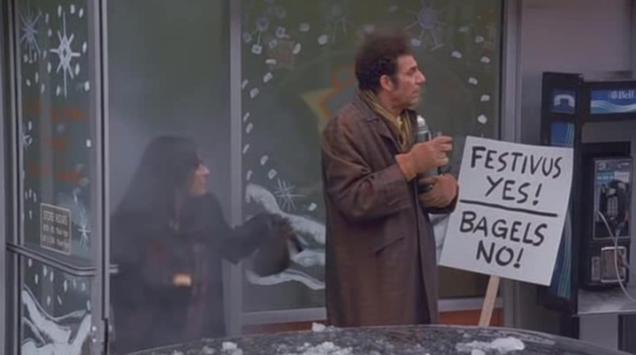 A man, resembling Kramer from 'Seinfeld,' holds a 'Festivus Yes! Bagels No!' sign outside a store with snowy decorations.