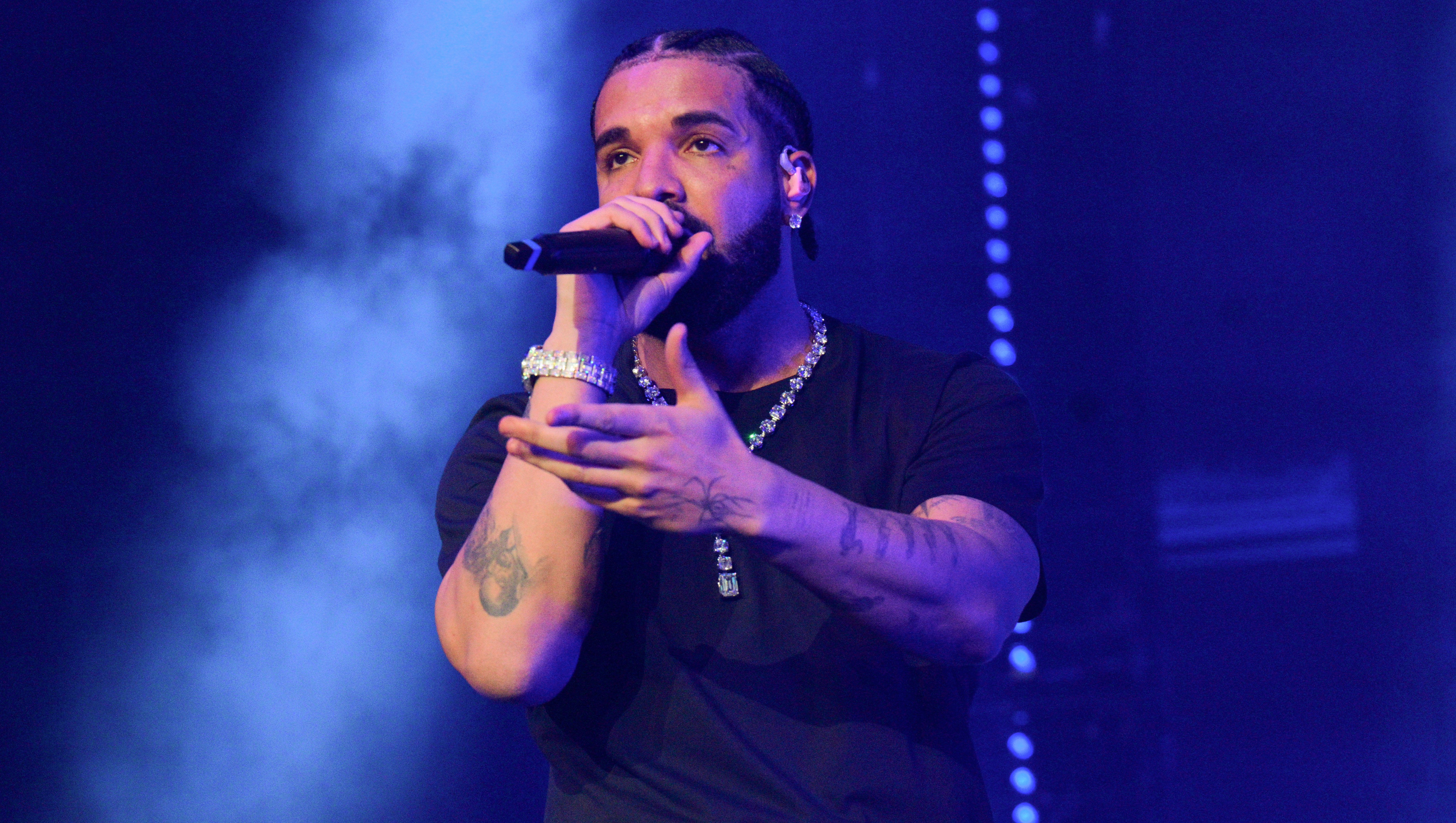 Drake in a black shirt performs on stage with a microphone, surrounded by blue lighting.