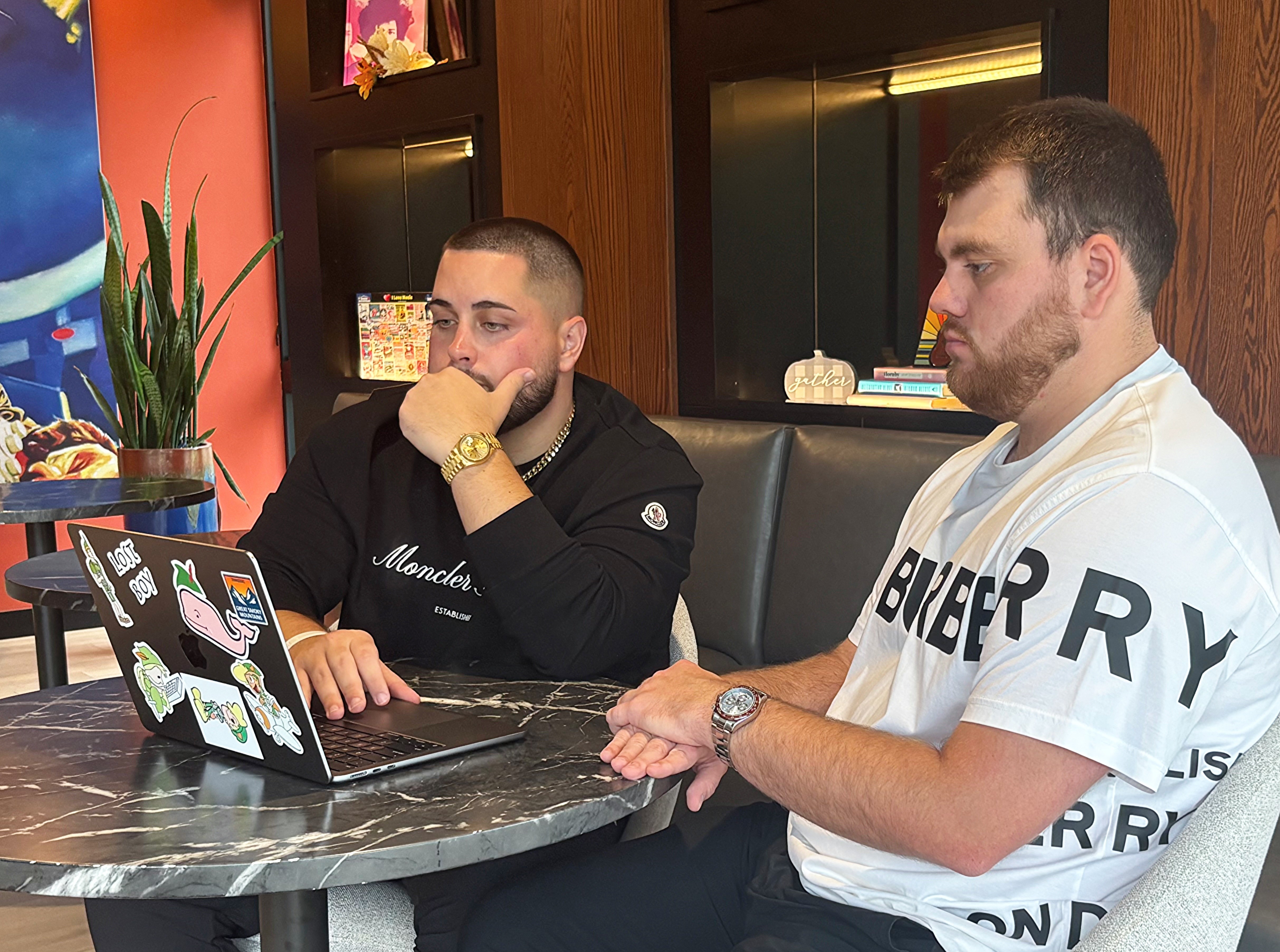 Two men sitting at a table, focused on a laptop covered with stickers. One wears a black Moncler shirt, the other a white Burberry shirt.