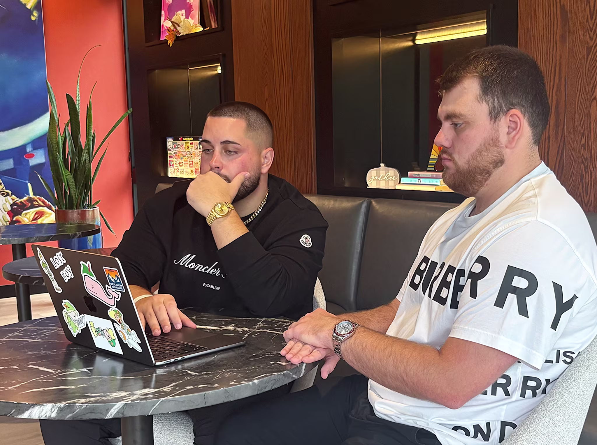 Two men sitting at a table, focused on a laptop with stickers. One wears a black Moncler sweatshirt, the other a white Burberry shirt.