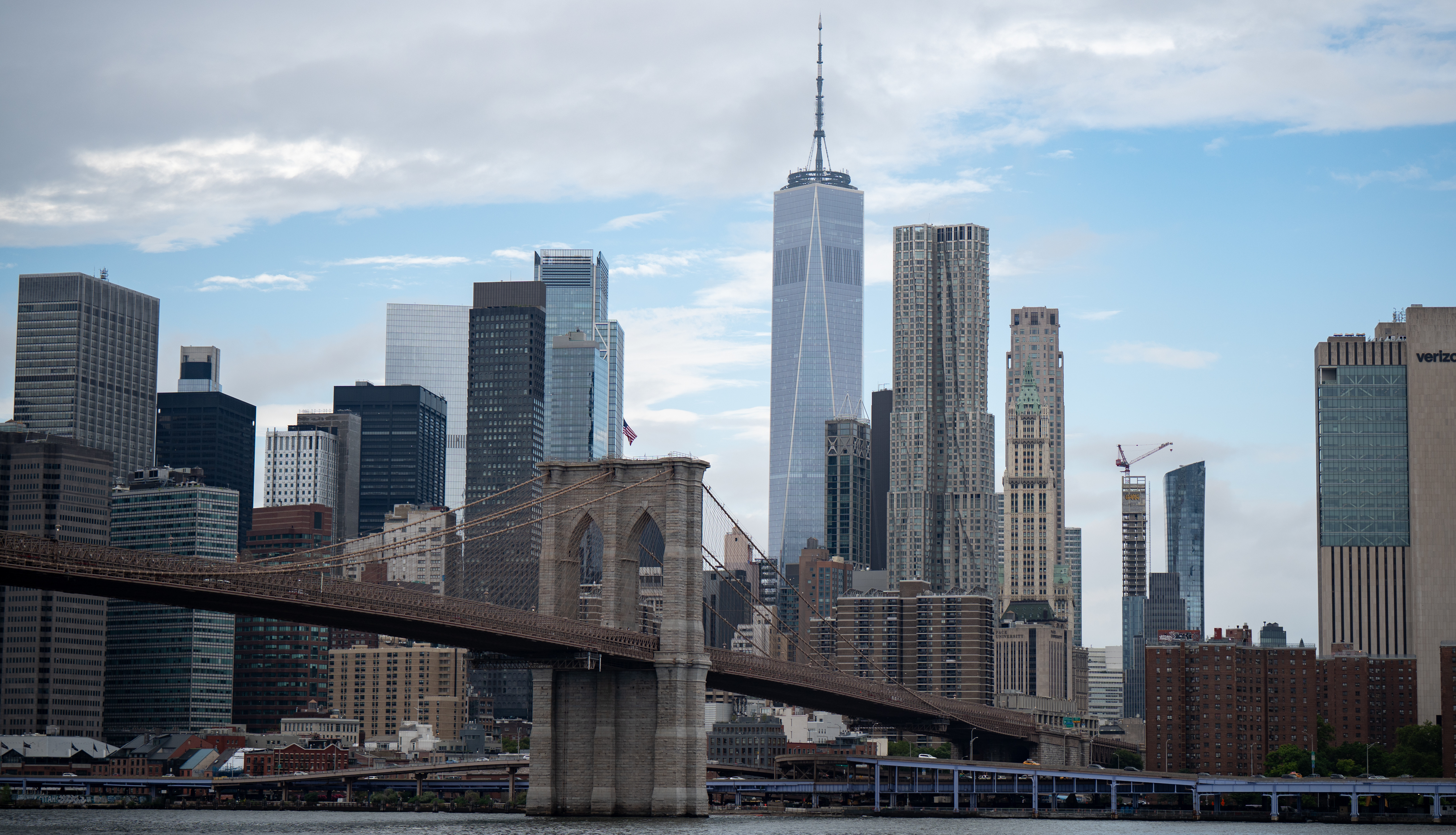 Brooklyn Bridge with New York City skyline, featuring One World Trade Center and various skyscrapers under a partly cloudy sky.