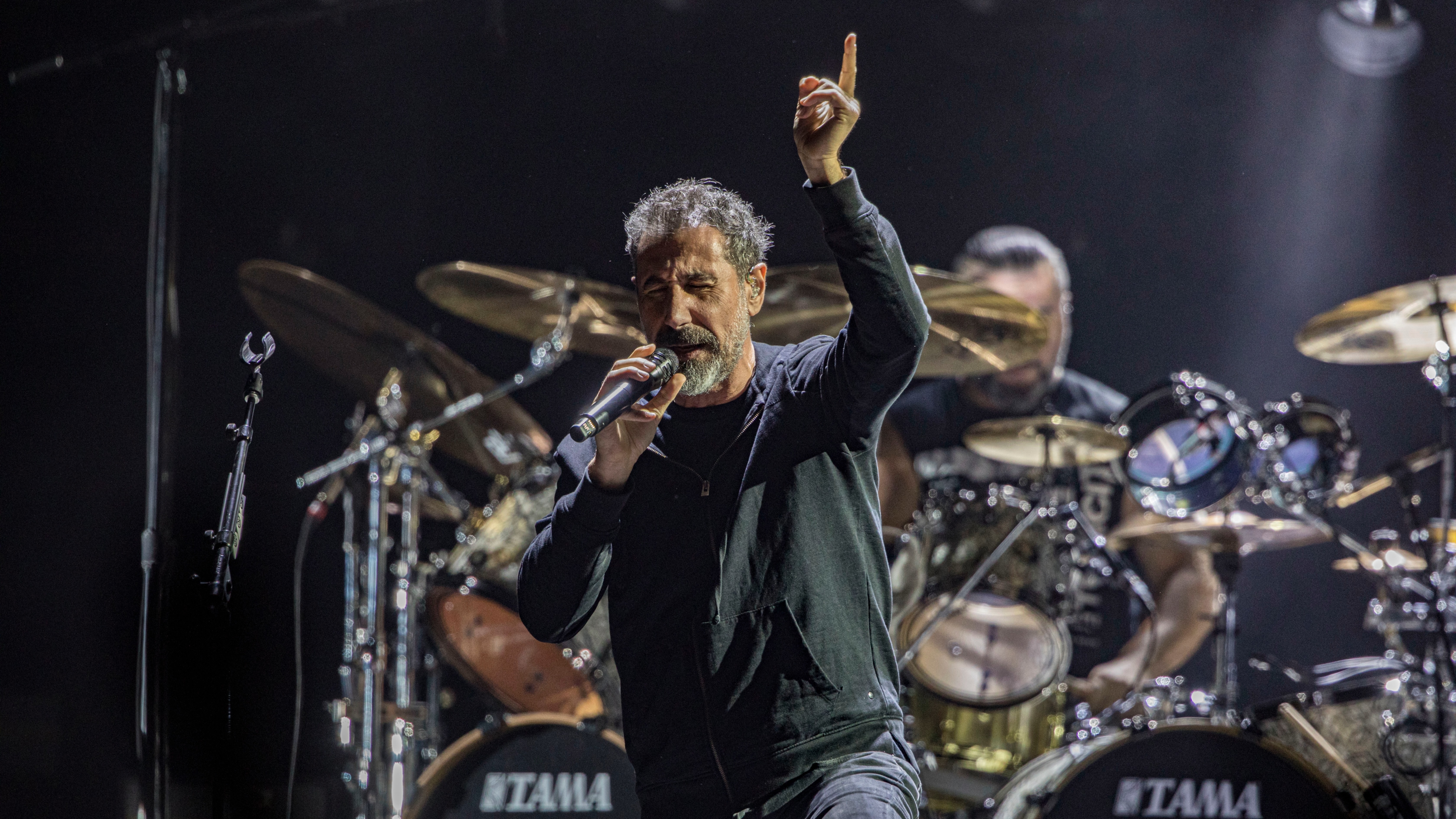 System of a Down lead singer on stage with a microphone, drummer in the background, surrounded by musical instruments and spotlights.
