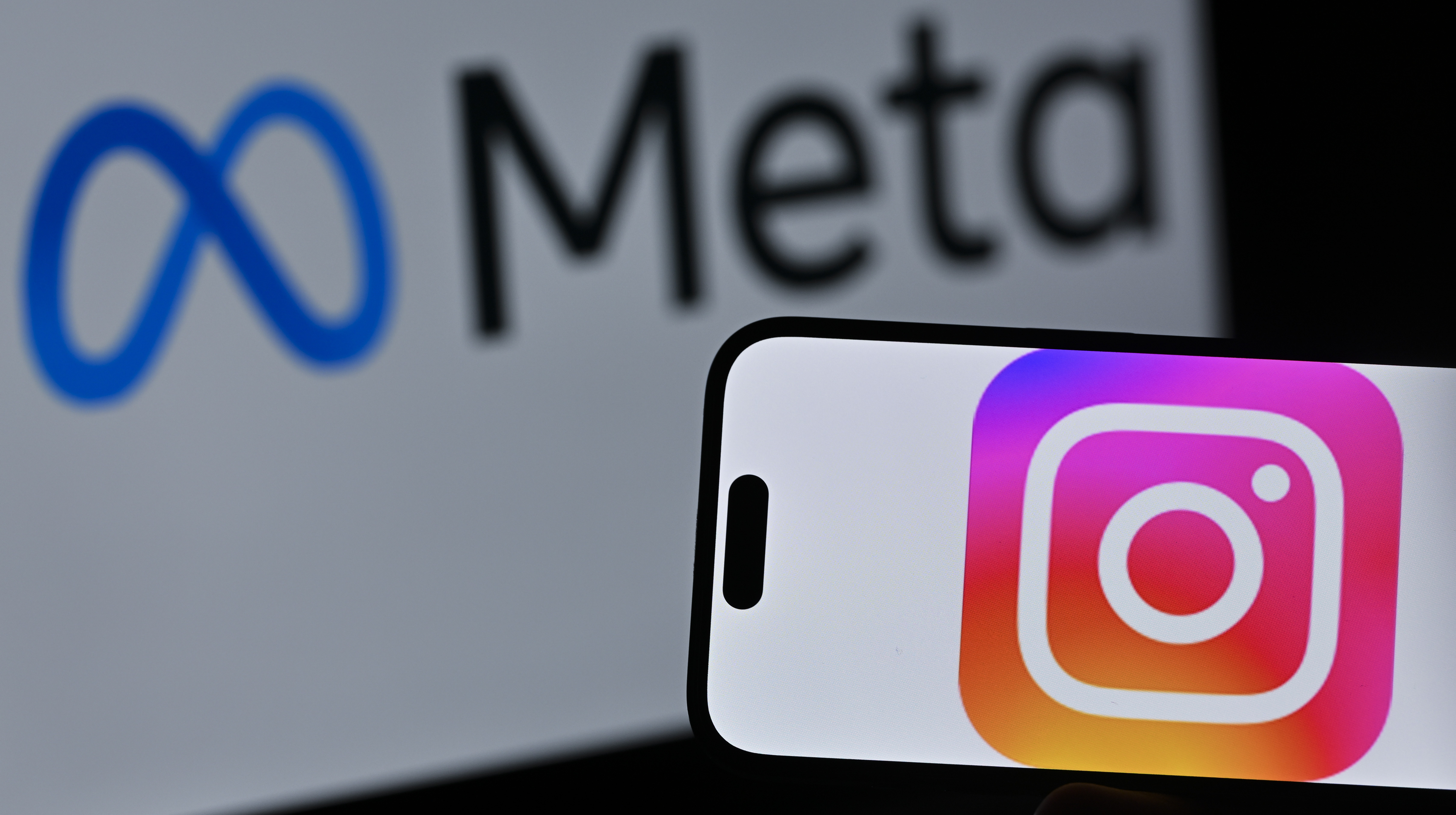 A smartphone displaying the Instagram logo is held up in front of a screen showing the Meta logo.