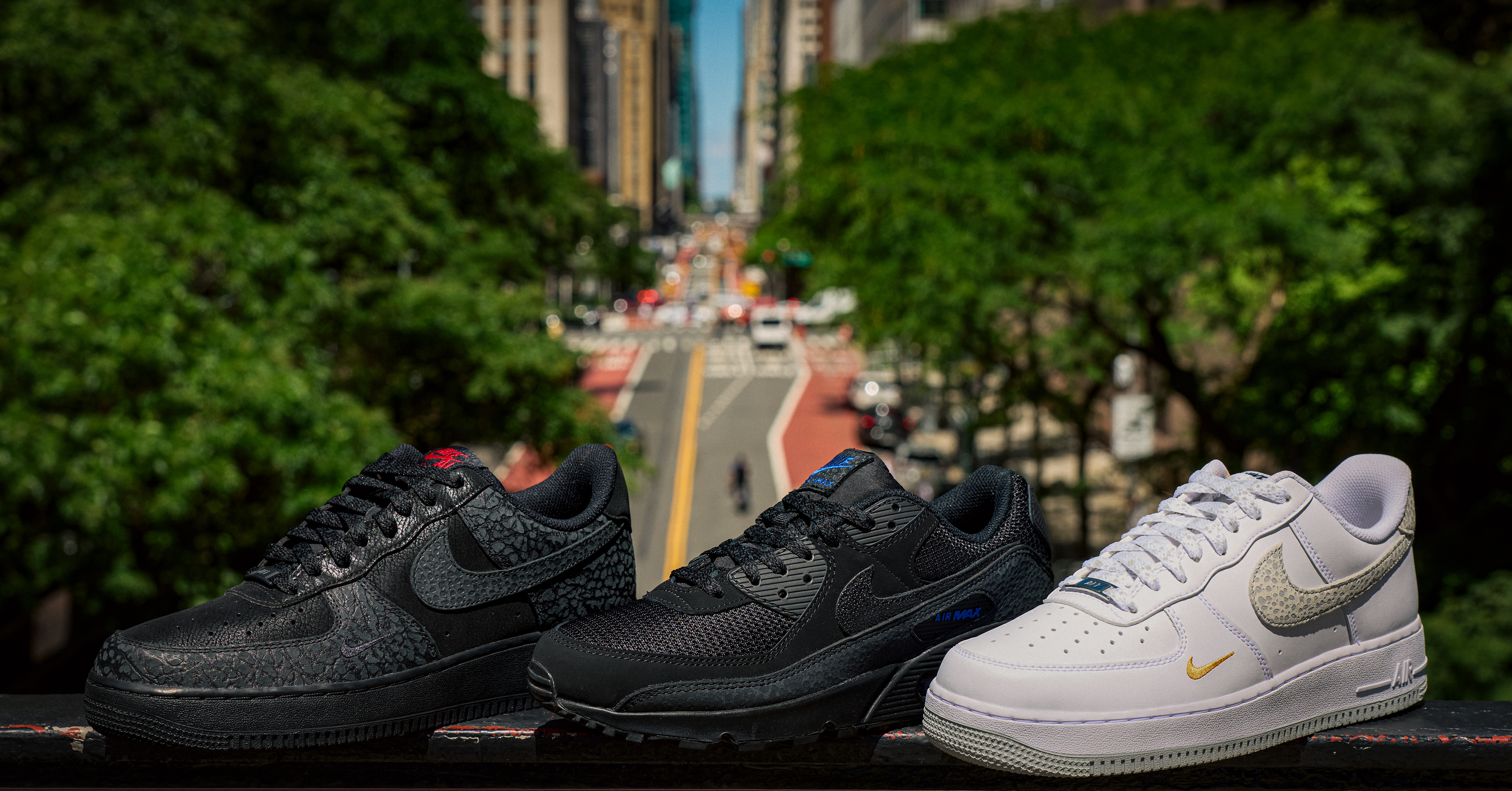Three pairs of Nike sneakers are displayed on a ledge with a blurred city street and greenery in the background.