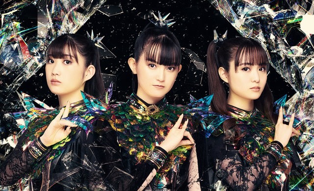 Three members of Babymetal in colorful, futuristic outfits with a dark, abstract background.