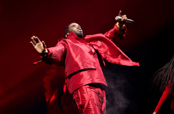Diddy on stage, holding a microphone, with dramatic lighting and smoke effects.