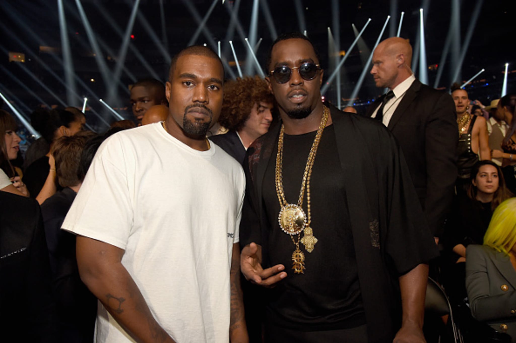 Kanye West and Sean "Diddy" Combs at an event, with Kanye in a white shirt and Diddy wearing sunglasses and a black outfit with chains.