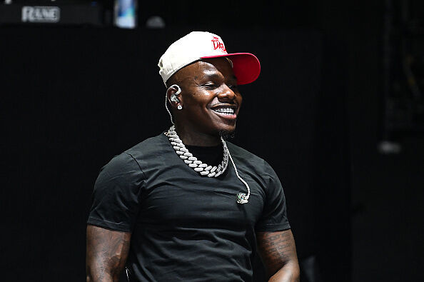 DaBaby, wearing a black shirt and white cap, smiles on stage. He has a diamond necklace and earpiece.
