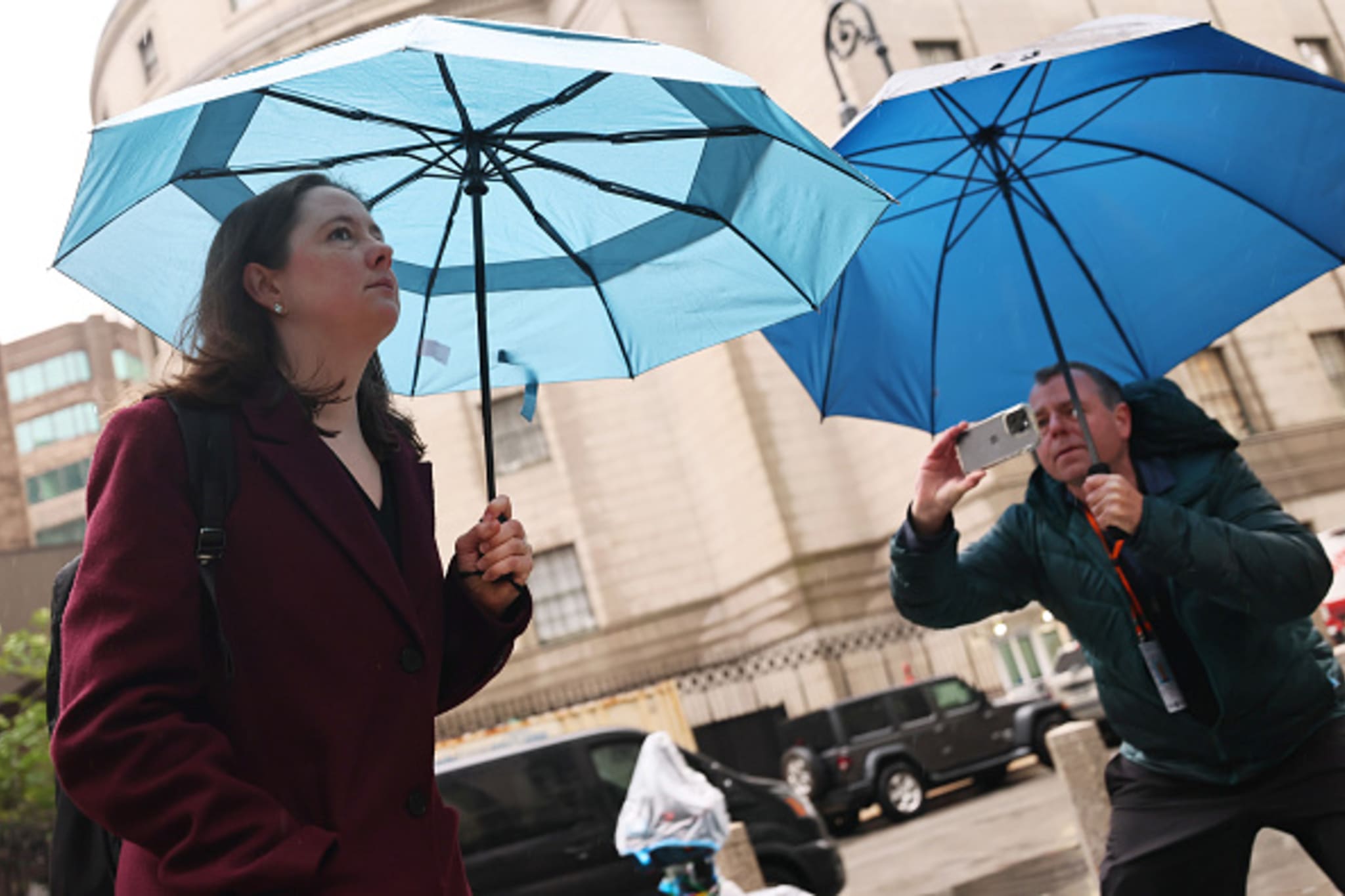 Two people with blue umbrellas on a city street; one takes a photo with a smartphone.