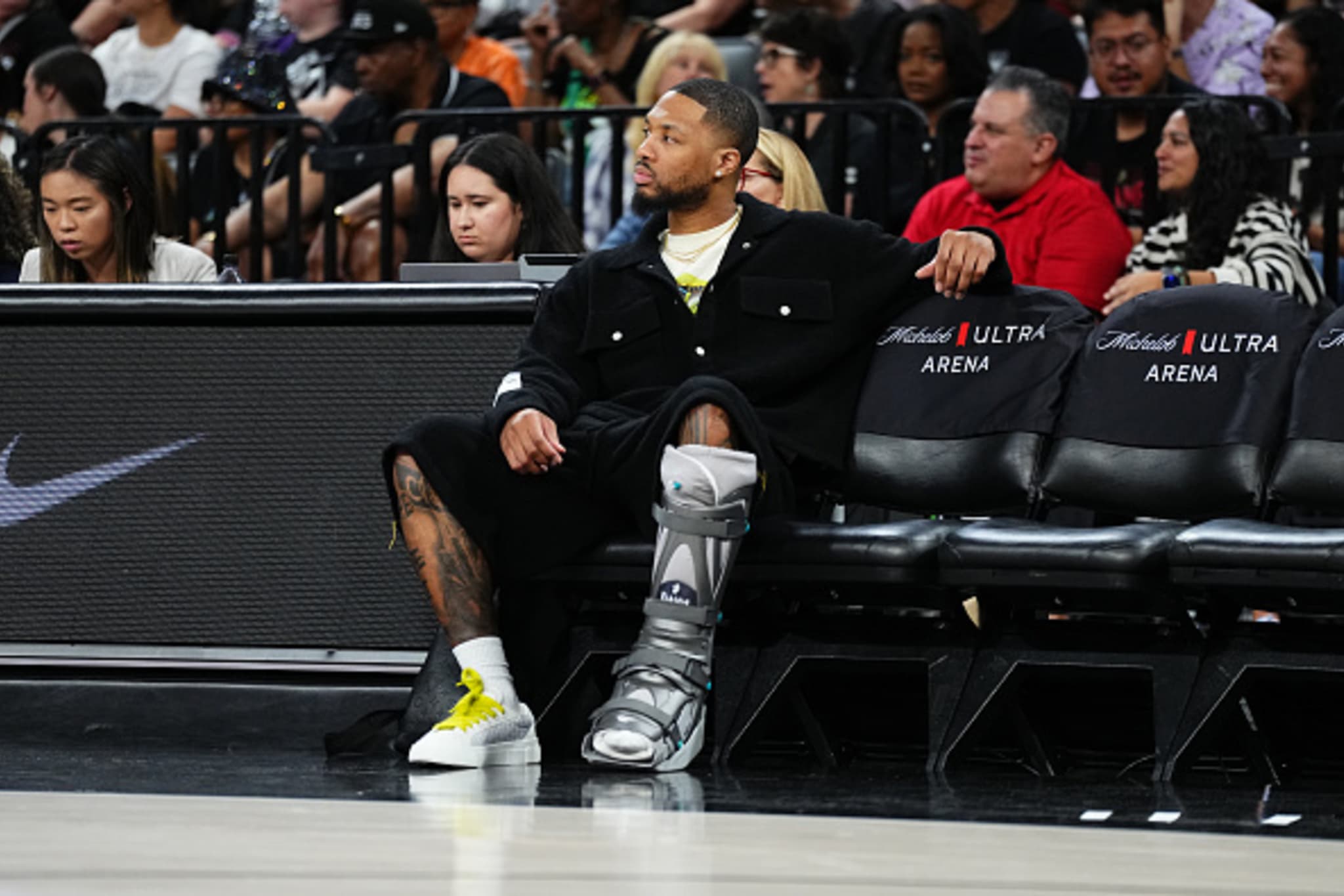 A man wearing a black outfit and a medical boot sits courtside at a basketball game, surrounded by spectators in an arena.