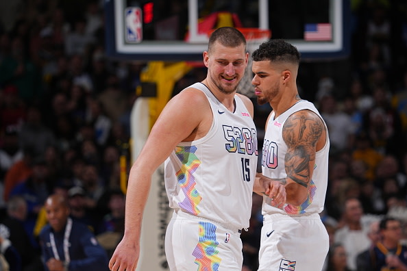 Michael Porter Jr. and Nikola Jokić in white jerseys with colorful accents on a court, interacting during a game.